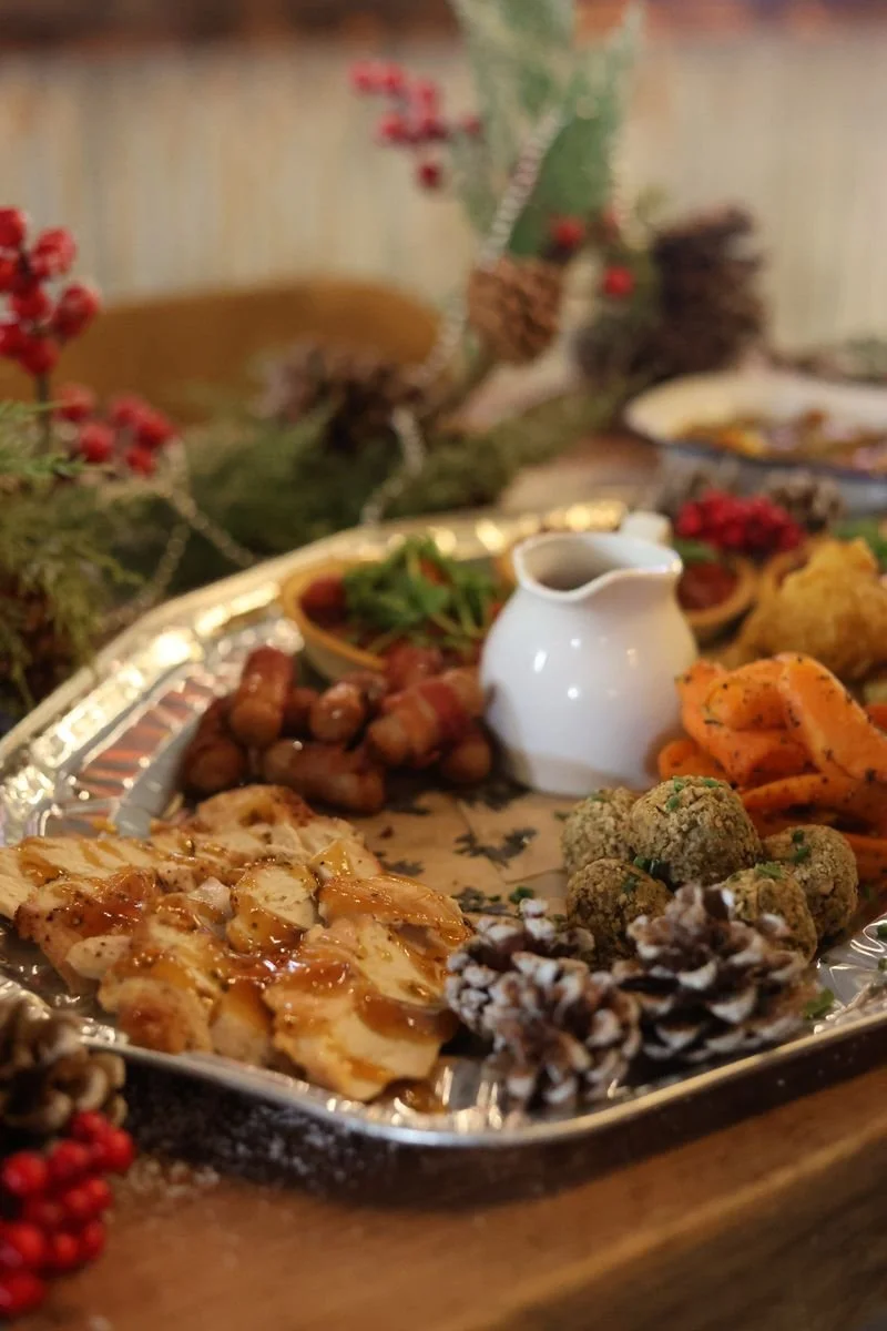 Festive holiday platter with glazed turkey slices, bacon-wrapped sausages, falafel balls, carrot ribbons, and a white gravy boat, surrounded by decorative pinecones and berries.