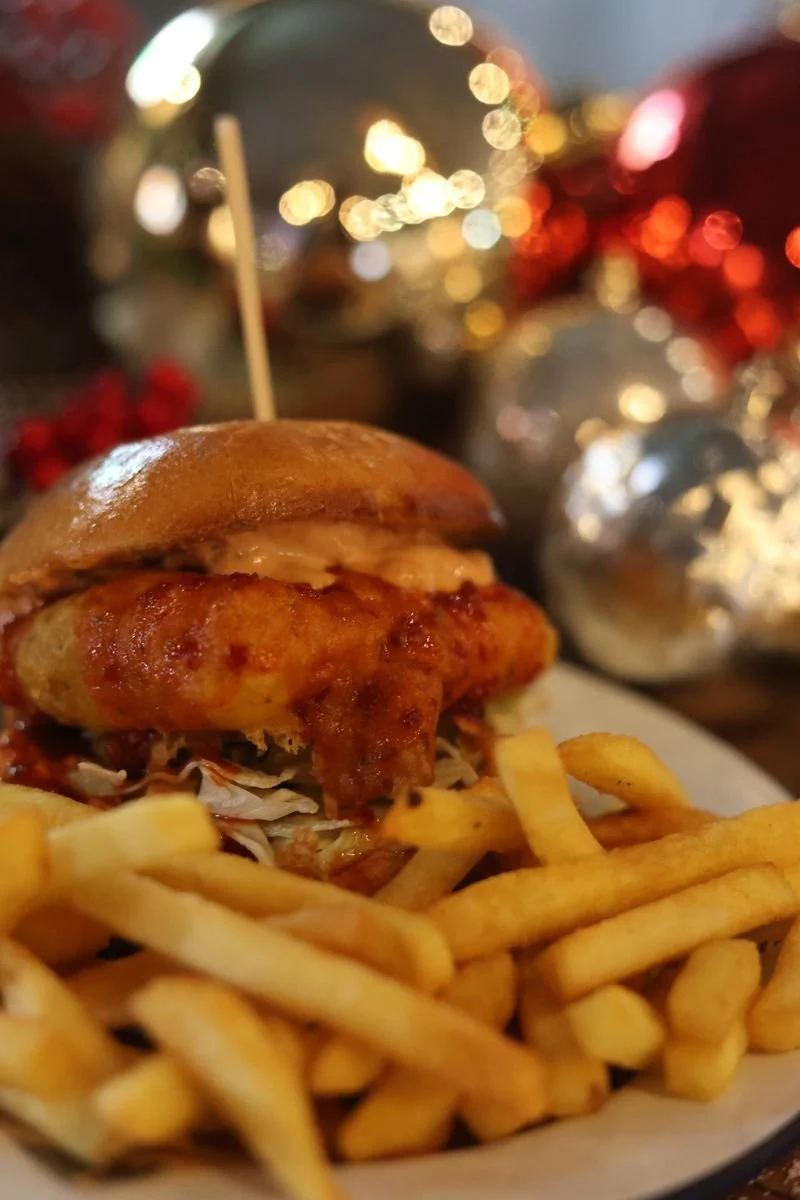 A breaded chicken burger with lettuce and sauce on a plate, accompanied by French fries. Blurred Christmas ornaments in the background.