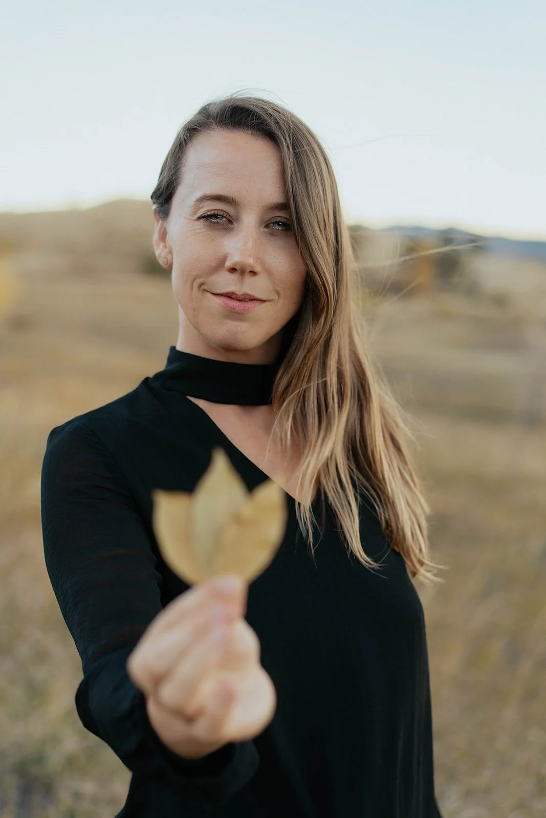 A woman with long hair wearing a black top holding a gold leaf-shaped award in an outdoor field during daytime.
