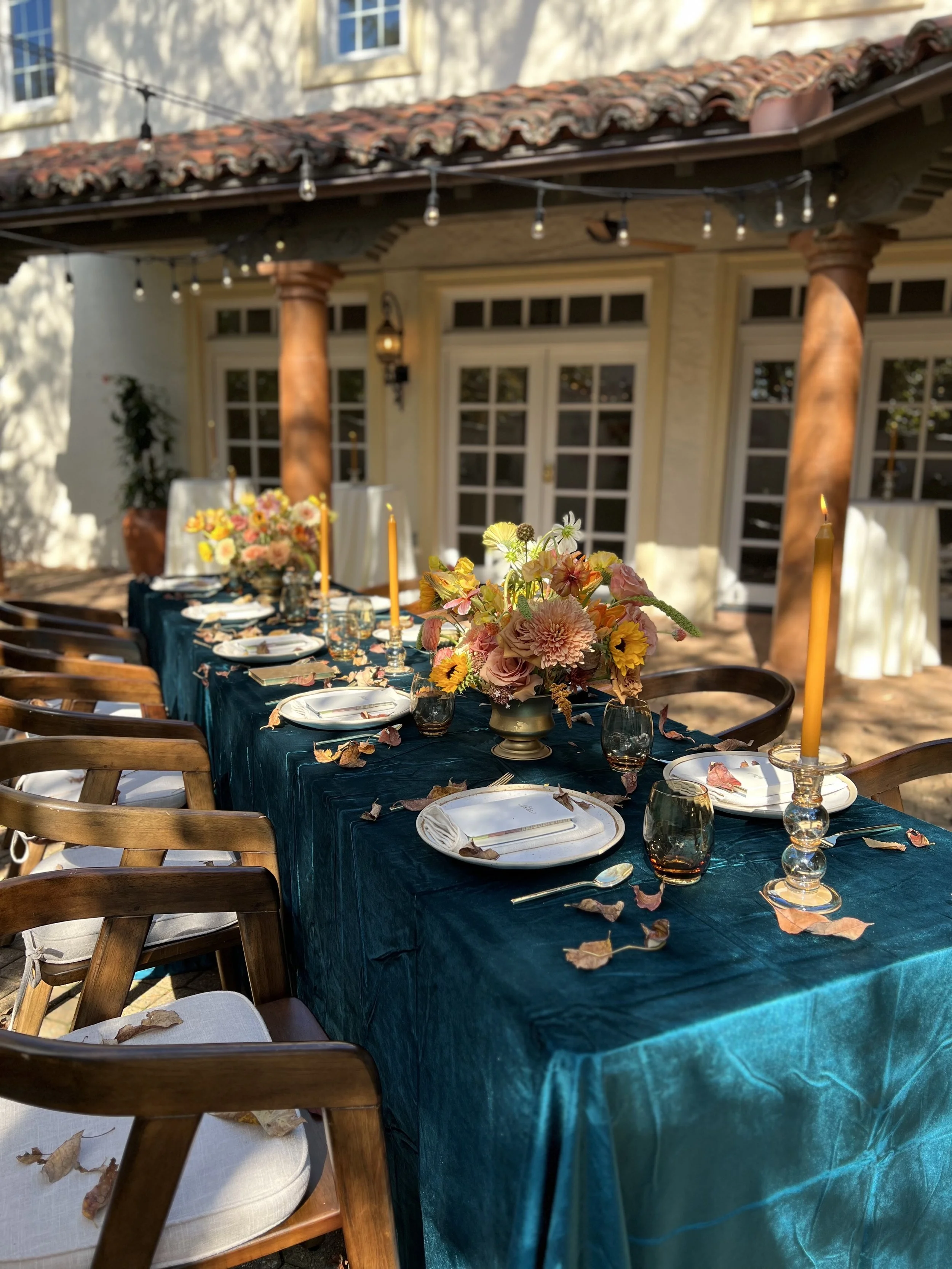 Outdoor dining table set for a meal with floral centerpieces, candles, and tableware, near a house with a tiled roof and string lights.