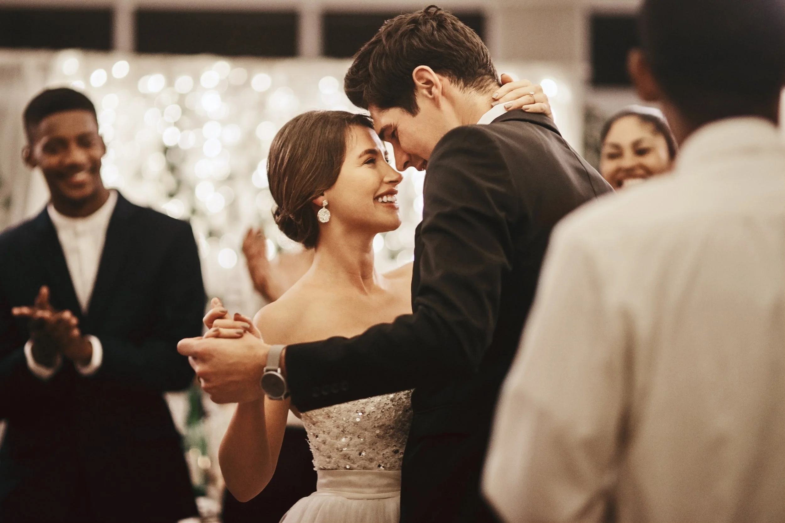 A young couple is dancing closely at a wedding reception, smiling and looking into each other's eyes, surrounded by happy guests.