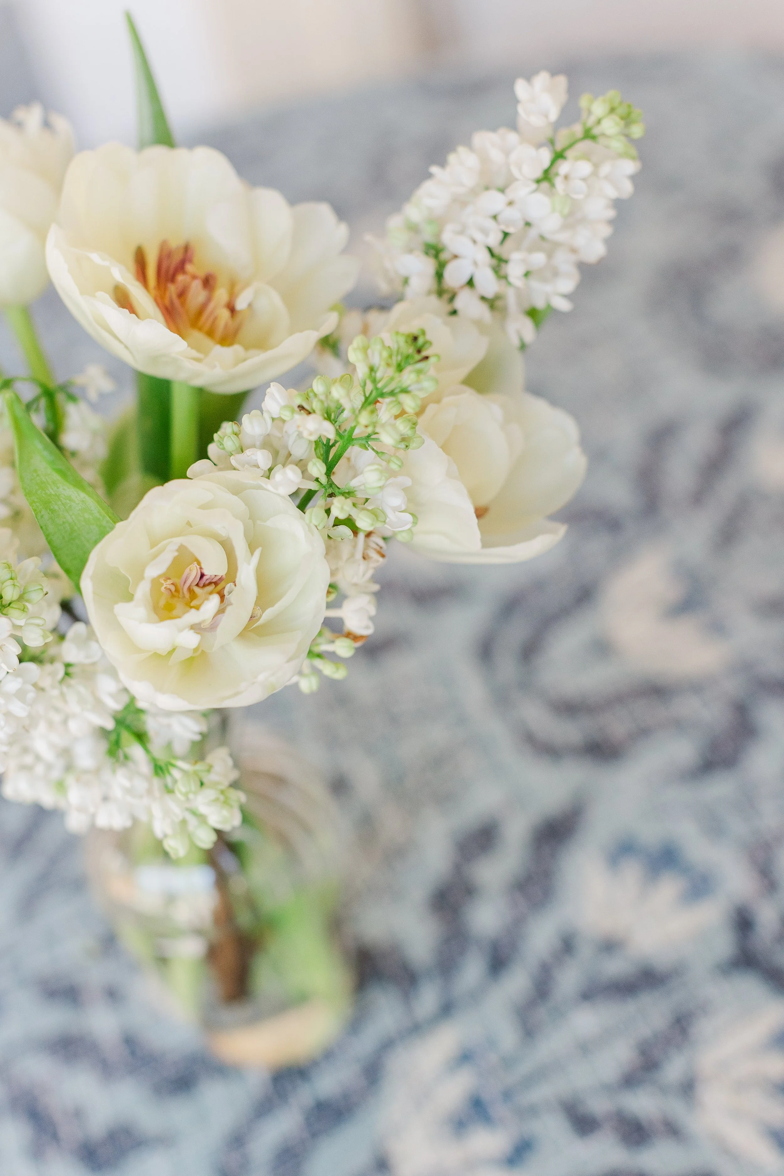 Close-up of a bouquet with white tulips, white lilacs, and greenery in a glass vase on a patterned table.