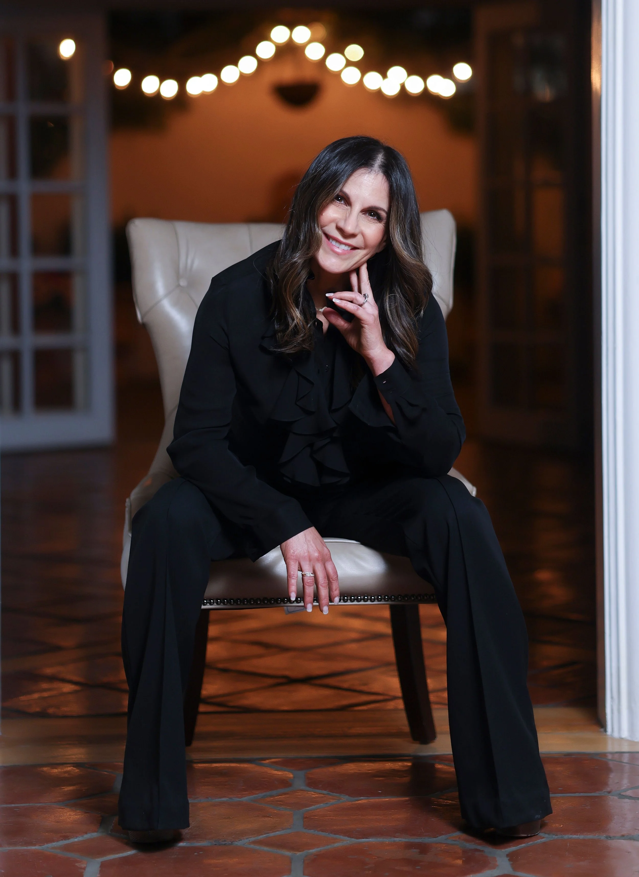 A woman with dark brown hair, wearing a black outfit, is sitting on a light-colored chair with a high back. She is smiling, resting her chin on her right hand, and looking at the camera. The background features string lights and a blurred indoor setting with warm lighting and tiled floor.