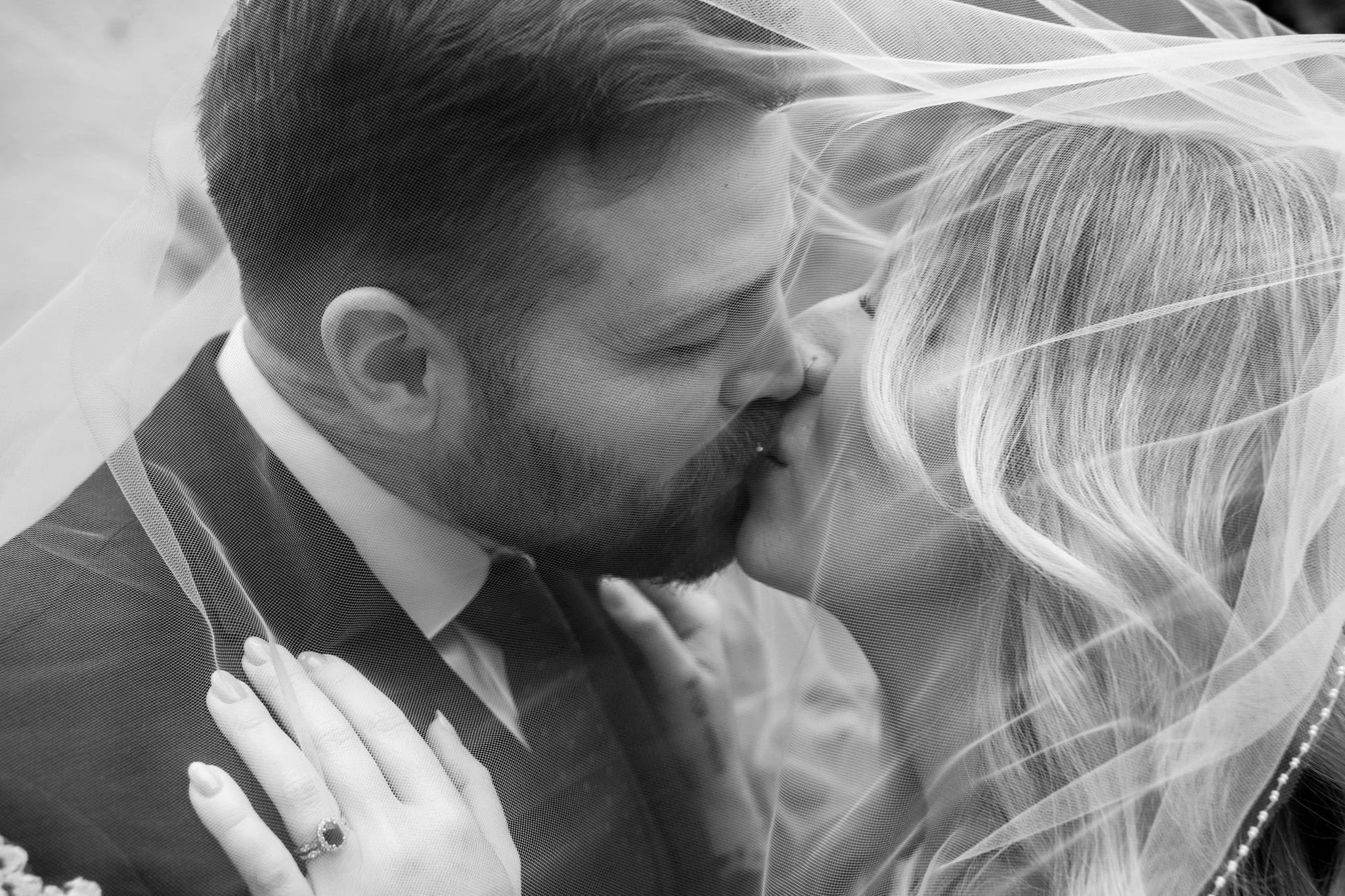 A black and white photo of a bride and groom kissing, with a veil partially covering their faces. The groom is wearing a suit and tie, and the bride has long, wavy hair and a ring on her finger.
