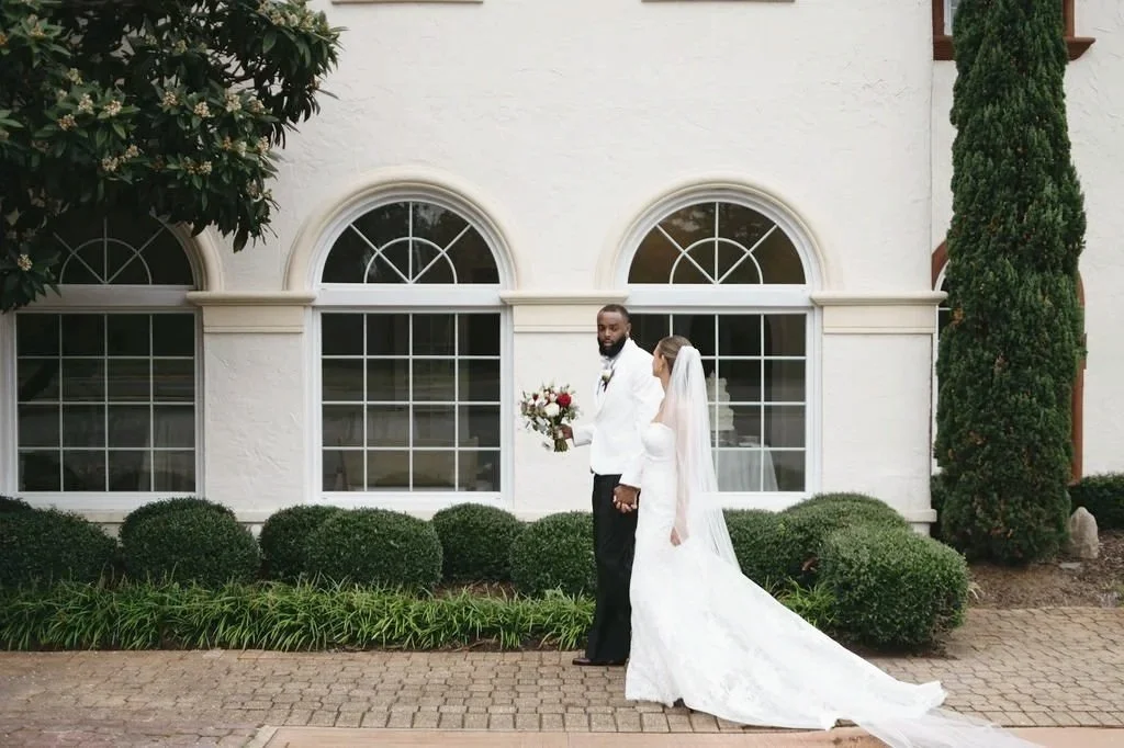 A bride and groom standing outside a white building with arched windows, the groom holding a bouquet of flowers, with greenery and bushes in the background.