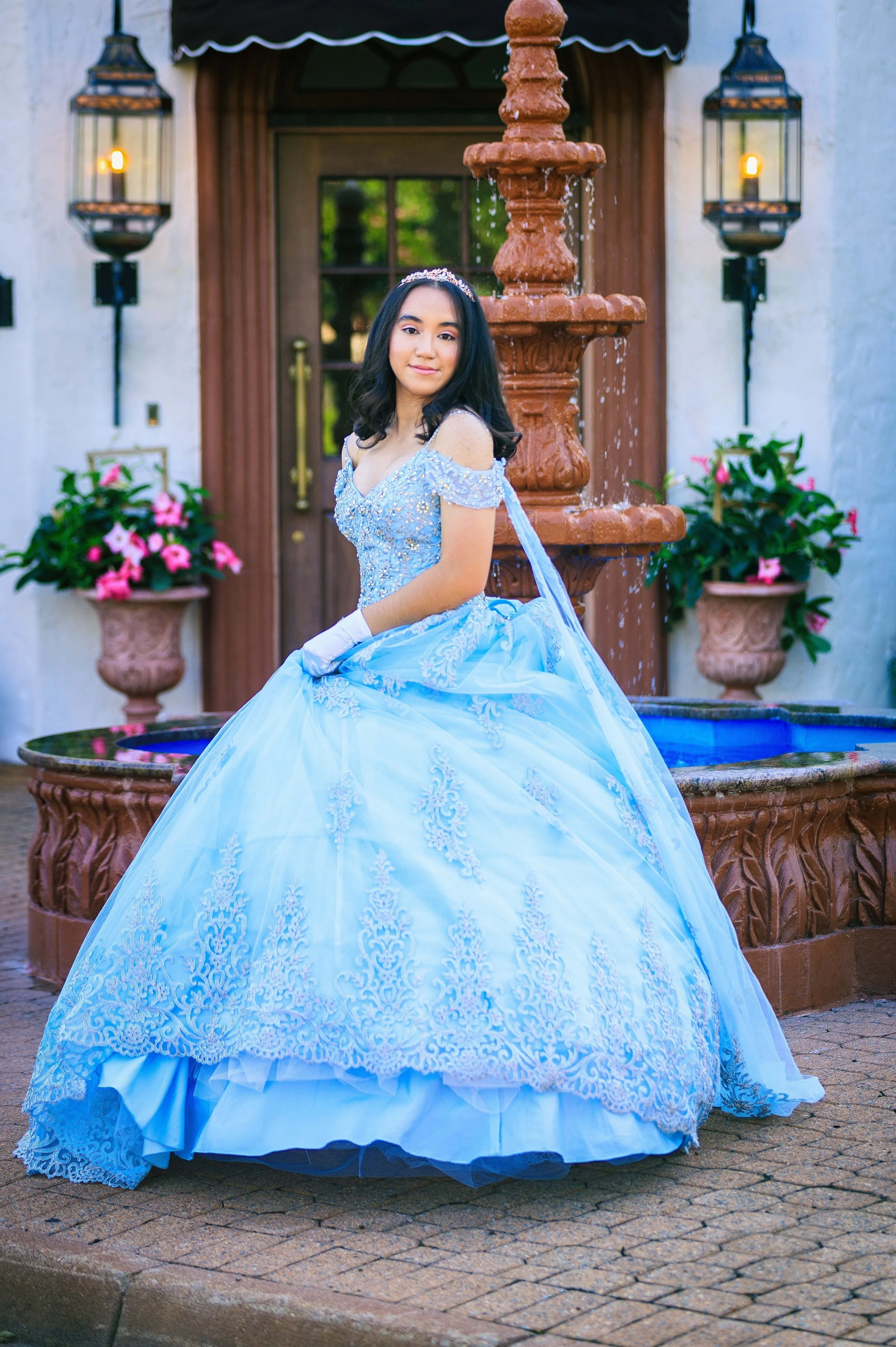 A young woman in a light blue princess dress with lace and bead embellishments, wearing white gloves and a tiara, sitting by a fountain in front of a building with lanterns and potted pink flowers.