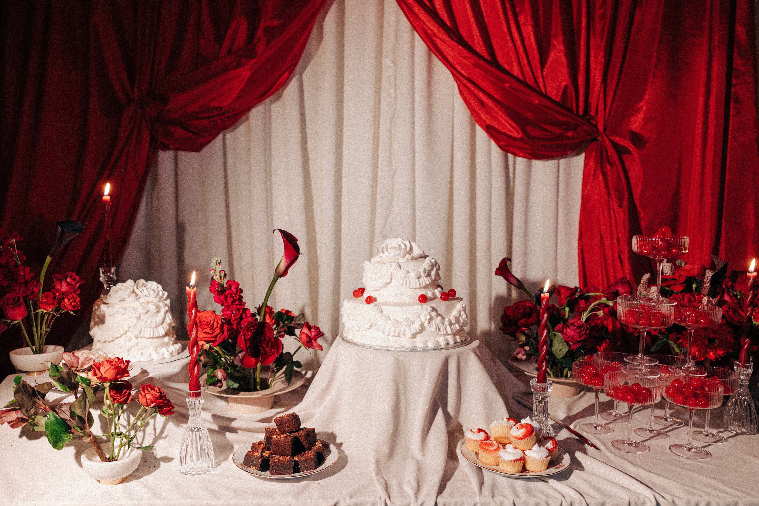 A dessert table decorated with red and white flowers, a tiered white cake with red berries, cupcakes, brownies, and glass bowls of cherries, set against red and white drapes