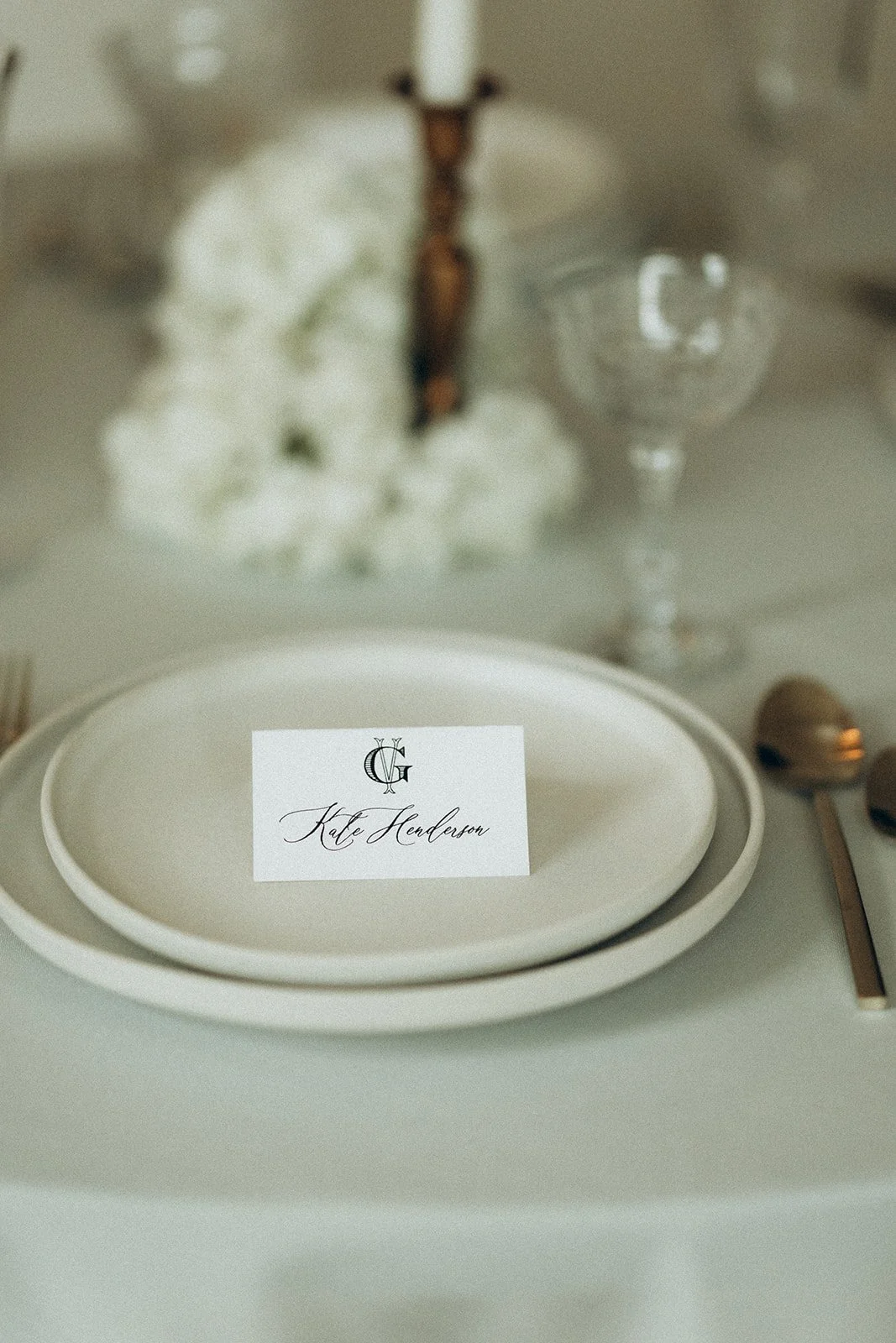 Elegant table setting with a name card reading 'Kate Henderson' placed on top of stacked white plates, with gold utensils and a glass in the background, and a floral arrangement with cotton flowers and a wooden decoration.