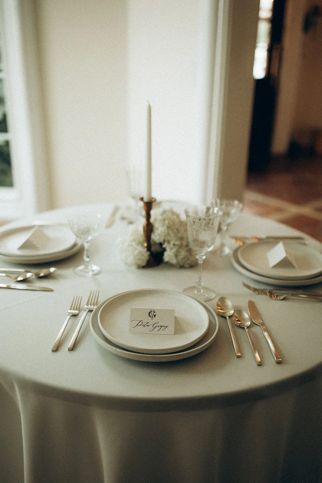 A round dining table set for a formal event with white plates, silverware, crystal glasses, and a candle centerpiece in a bright, elegant room.
