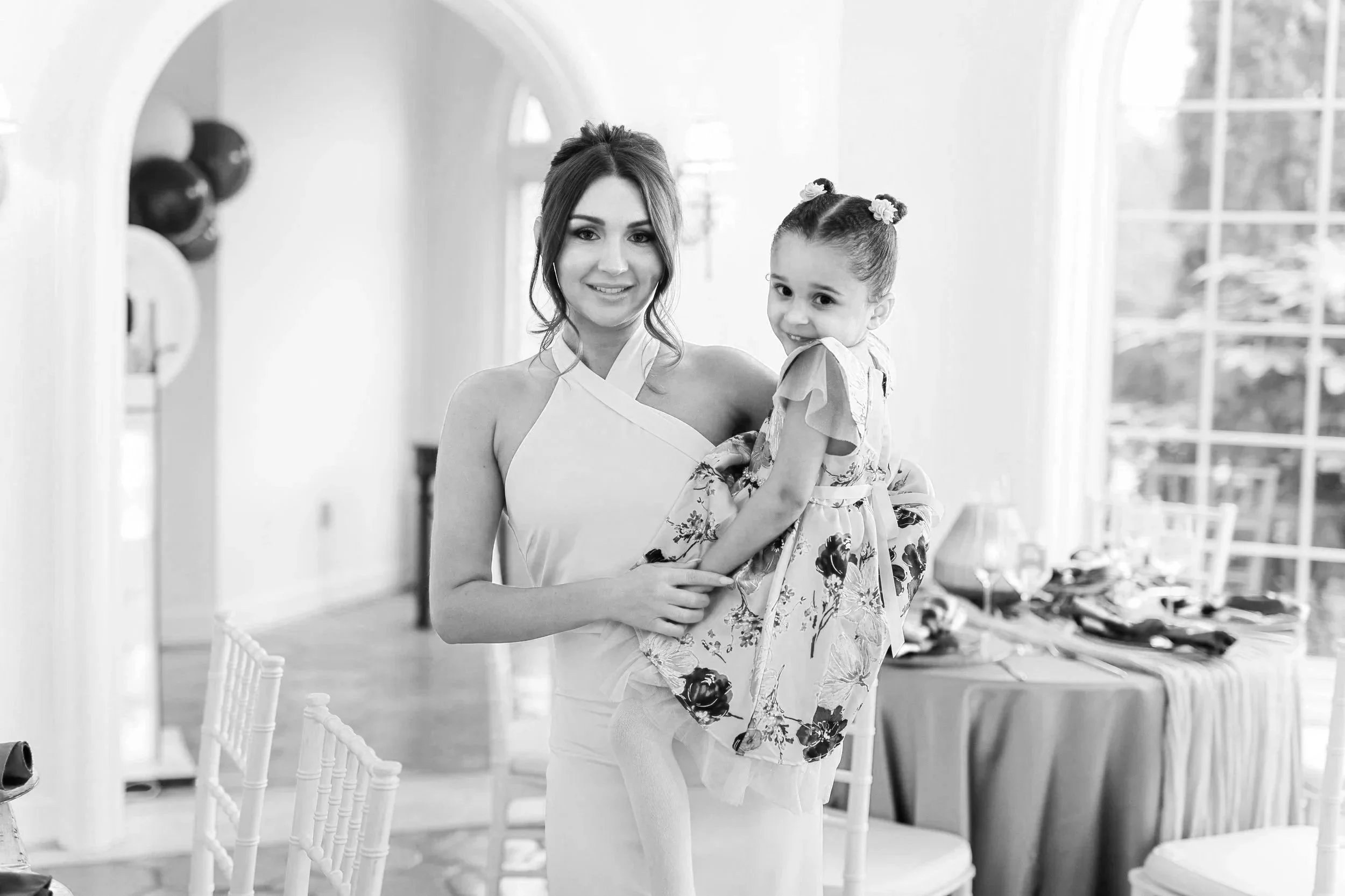 A woman holding a young girl inside a bright room with large windows and elegant decor.