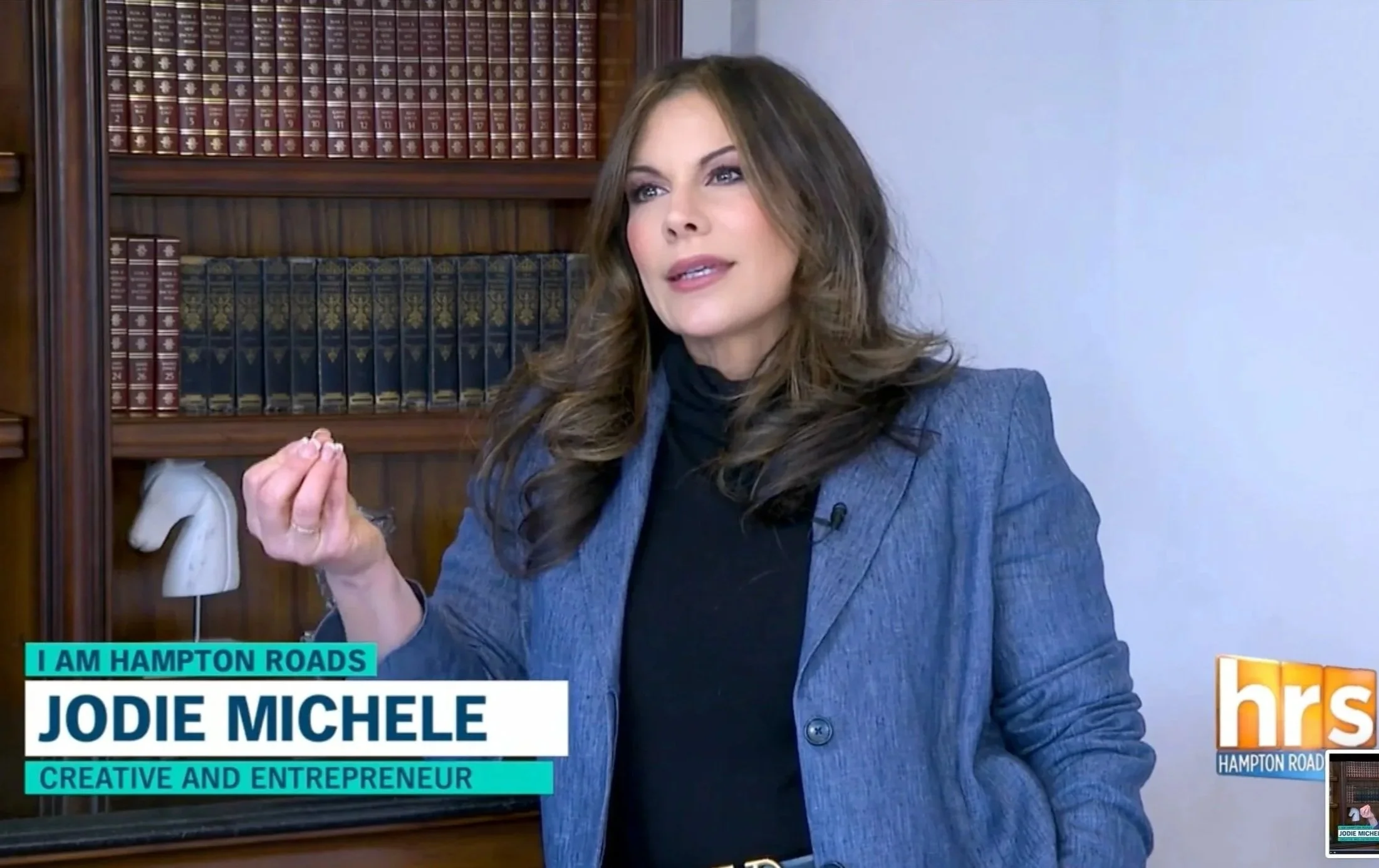 Jodie Michele wears a TWP suit while talking during an interview with WAVY TV 10 and Patrice Covington in a room with a bookshelf filled with hardcover books and a white horse-shaped ornament in the background.
