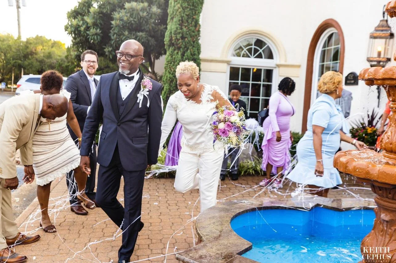 A joyful wedding celebration outside a building features a couple walking by a fountain, surrounded by guests. The groom wears a black tuxedo with a bow tie, and the bride wears a white outfit with a bouquet of purple and white flowers. Guests are dressed in colorful attire, some are holding streamers, and a few are bending down towards the fountain.