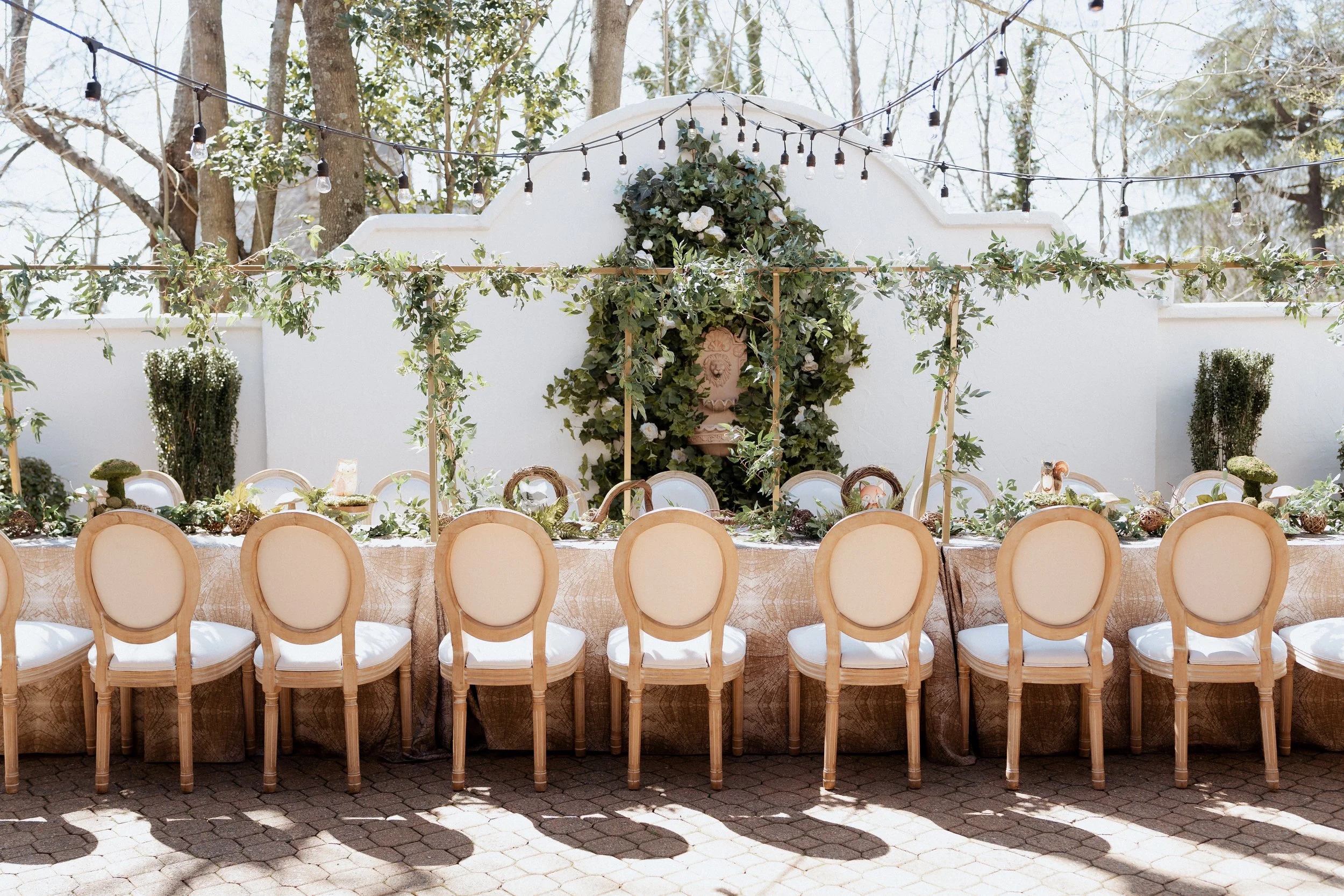 Outdoor wedding reception setup with a long table, cream-colored chairs, greenery, string lights, and floral decorations.