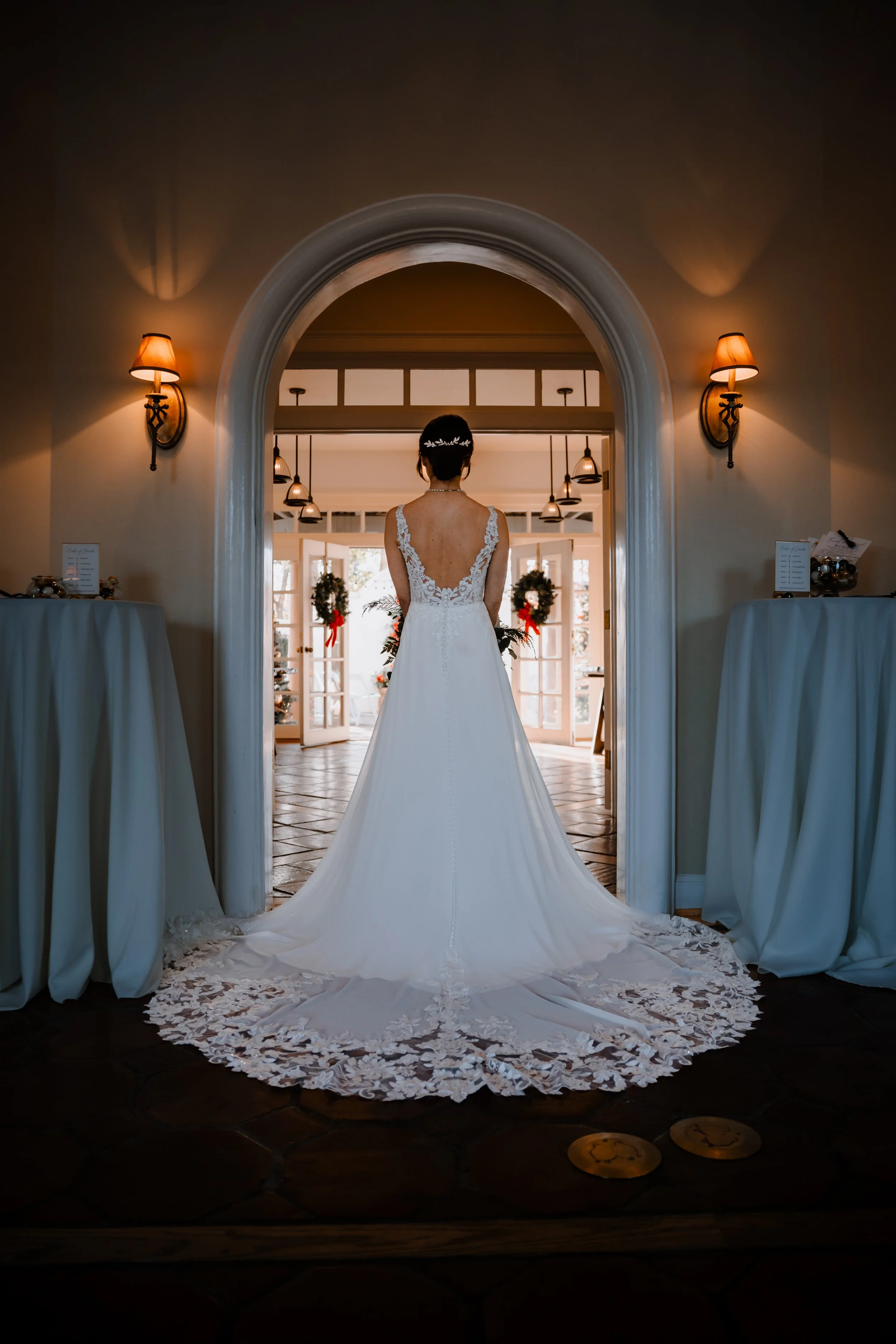 A bride in a white wedding gown standing in an arched doorway, looking into a warmly lit room decorated for the holidays with wreaths and red ribbons.