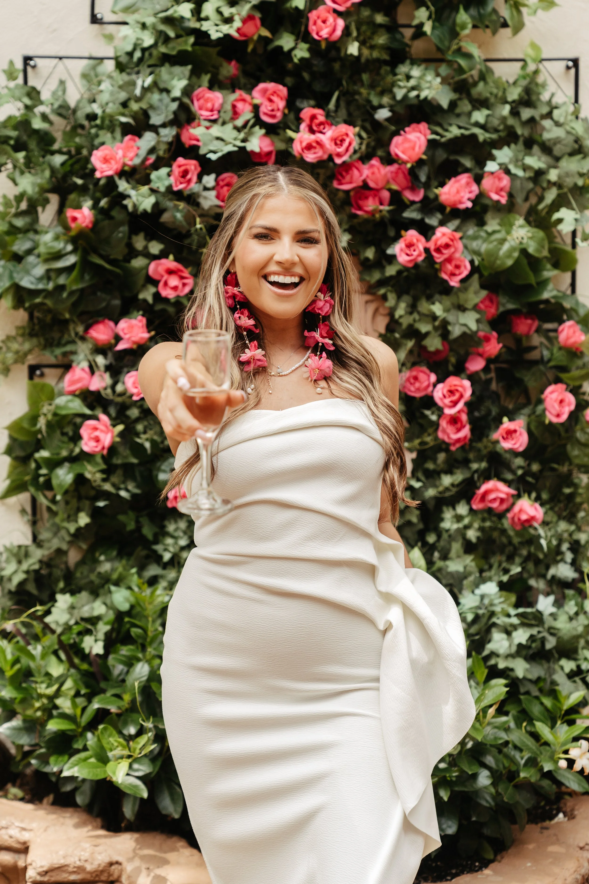 A woman in a white strapless dress with pink floral earrings and necklace is smiling and offering a wine glass, standing in front of a pink and green floral wall.