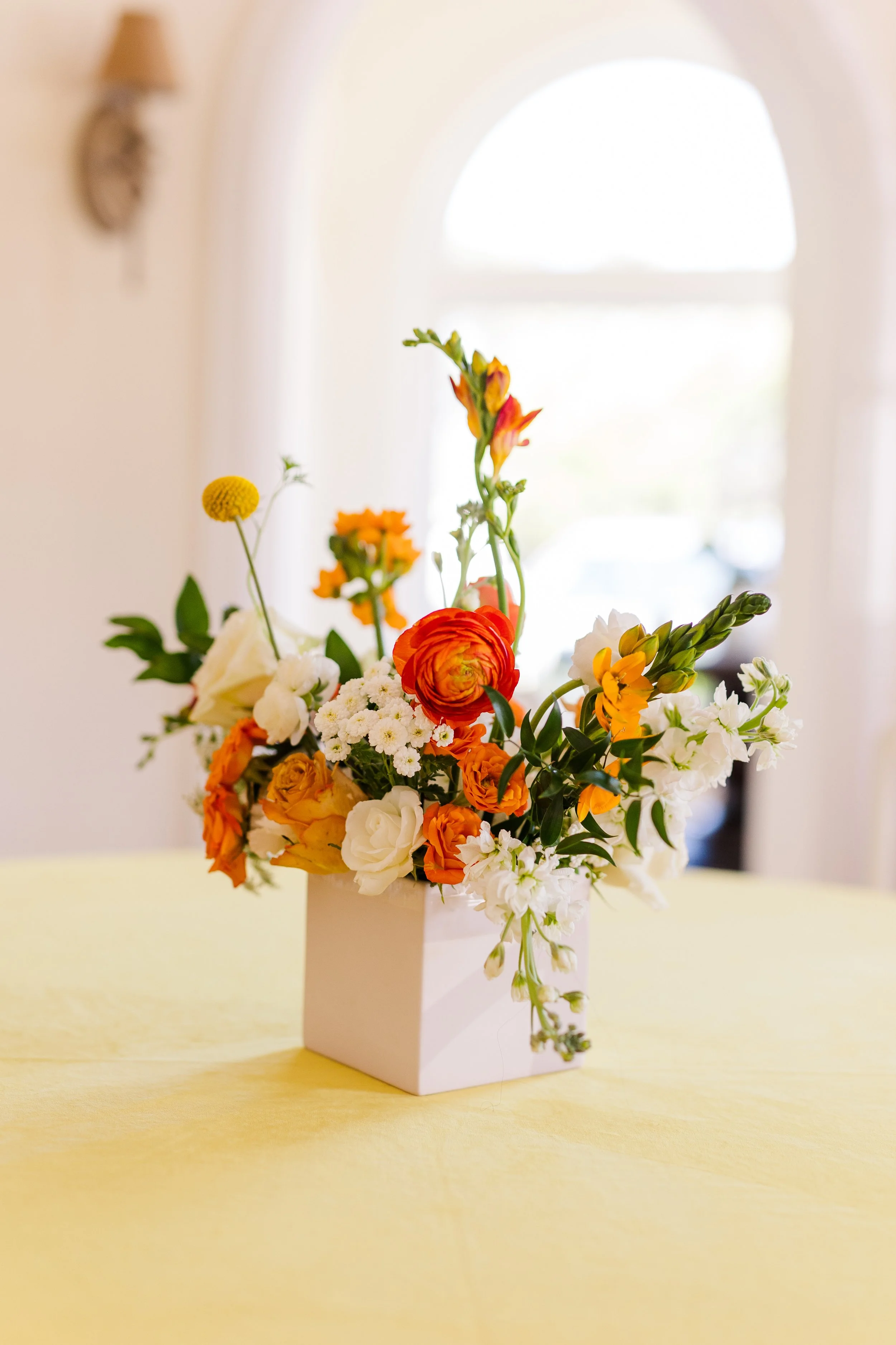 Colorful flower arrangement in a white vase on a yellow table, with a bright window and hanging light in the background.