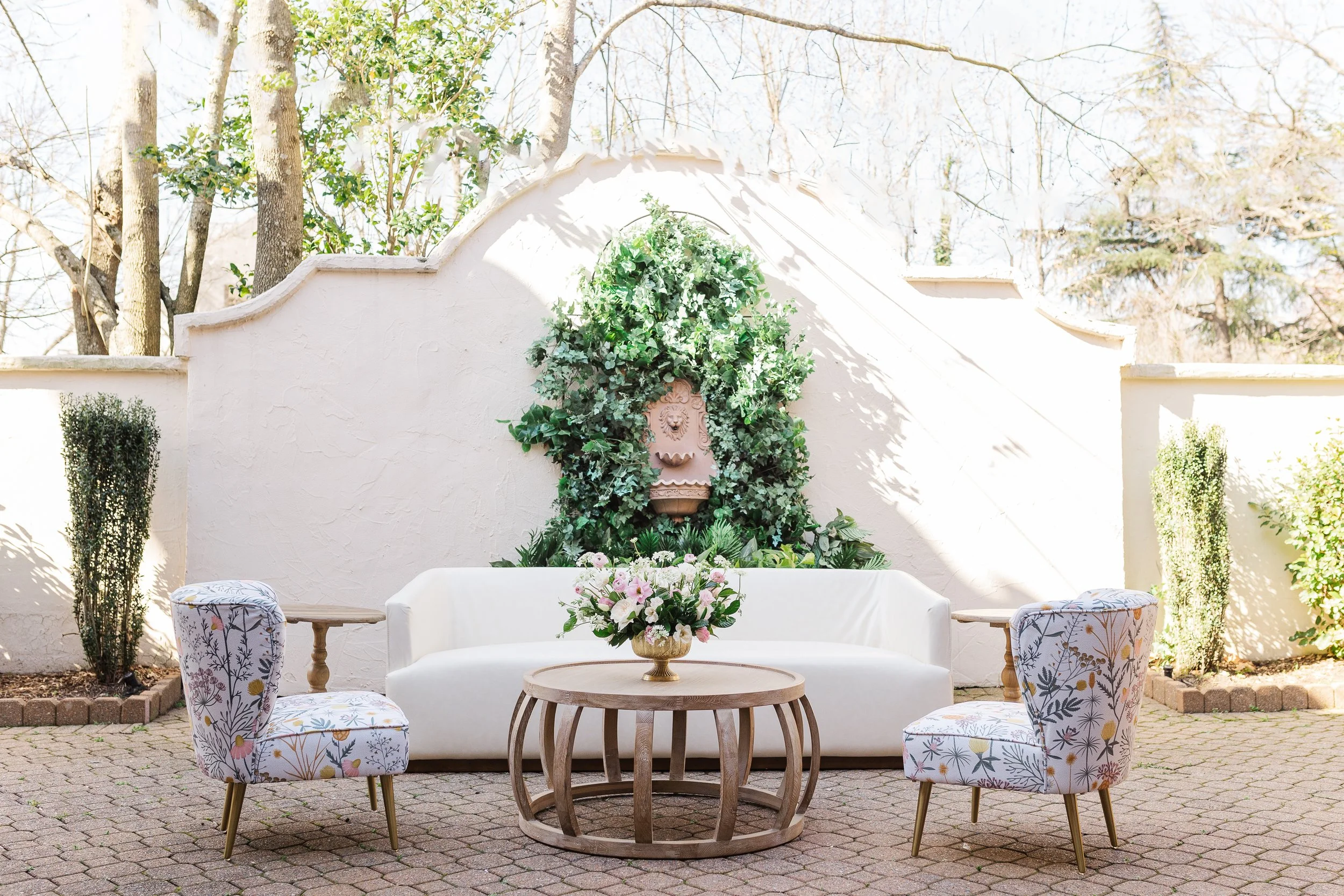 Outdoor patio seating area with a white sofa, two floral armchairs, a round wooden coffee table with a flower arrangement, and a decorative wall fountain with greenery in the background.