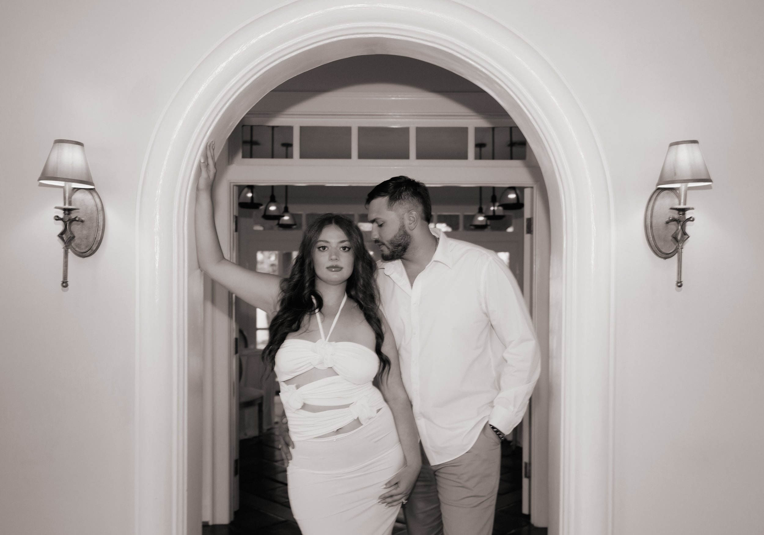 A black and white photo of a woman in a strapless dress with a knot at the front and cutouts, standing in a doorway with a man in a white shirt and light-colored pants. The woman has long, curly hair, and the man leans in toward her, with his hand in his pocket.