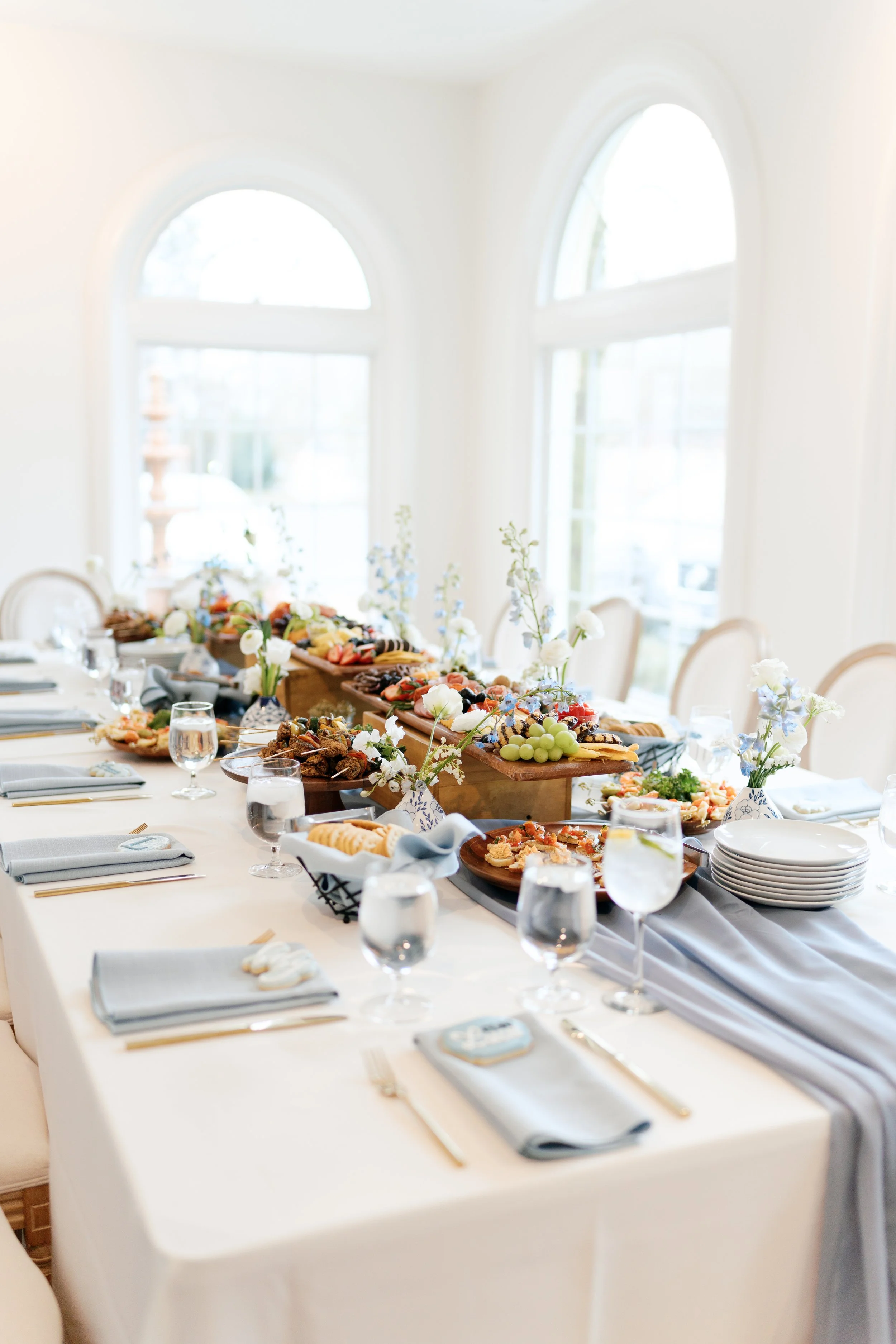 A decorated dining table set with plates, glasses, napkins, and a variety of food including fruits, appetizers, and desserts, with floral centerpieces.