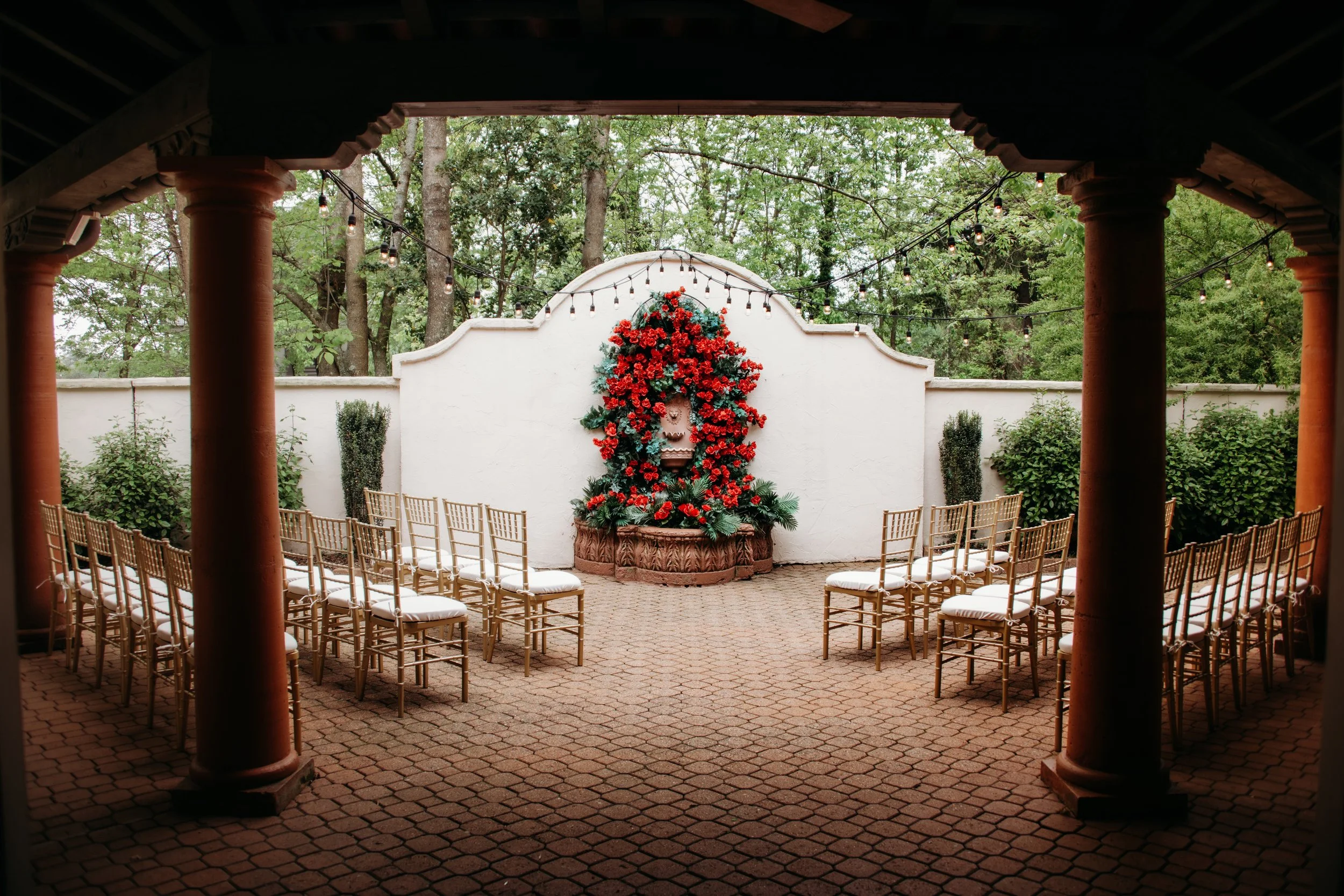Wedding ceremony setup with rows of chairs facing a white wall decorated with red flowers and greenery, outdoor setting with trees and string lights.