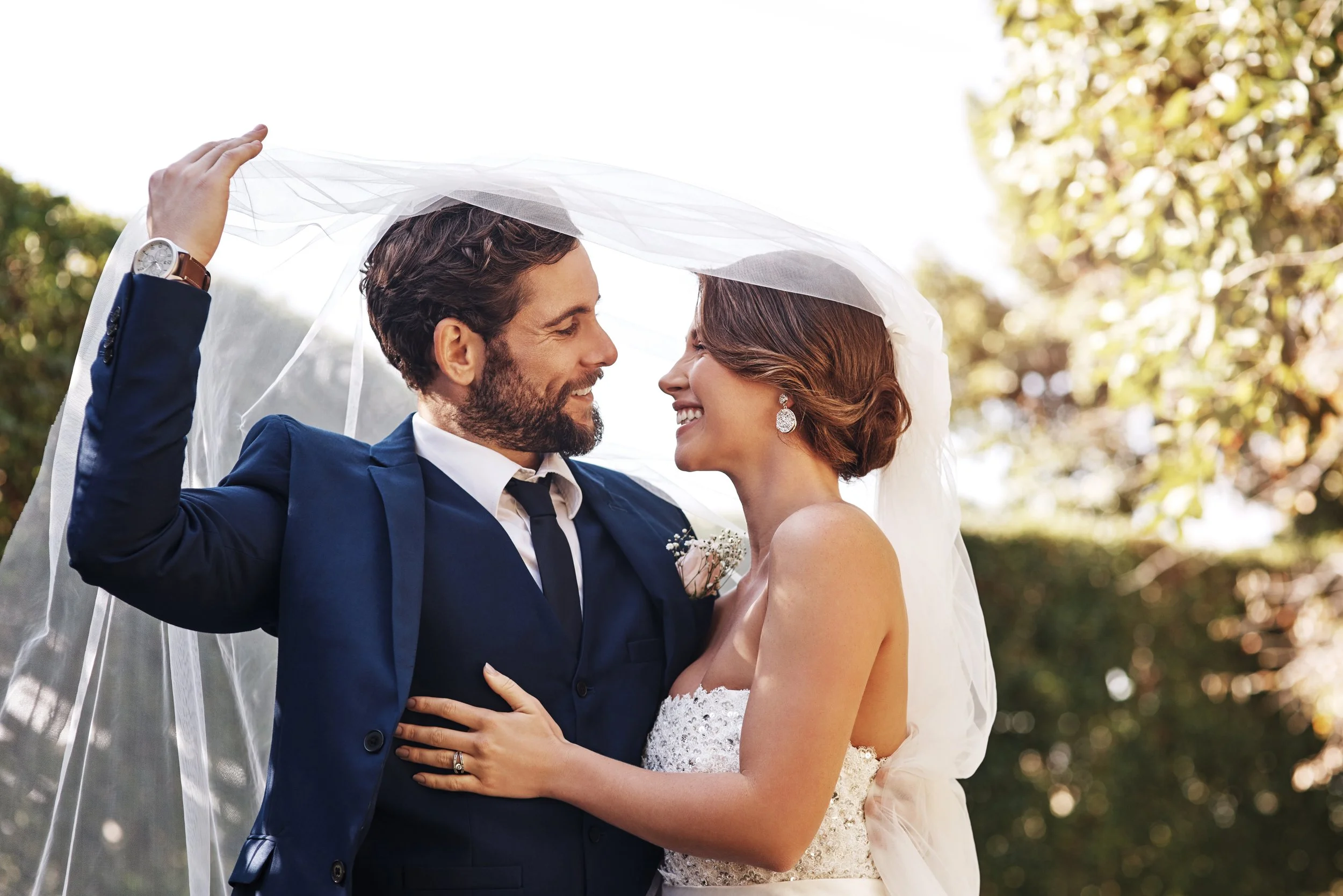 A newlywed couple smiling and looking at each other under a wedding veil outdoors with trees in the background.