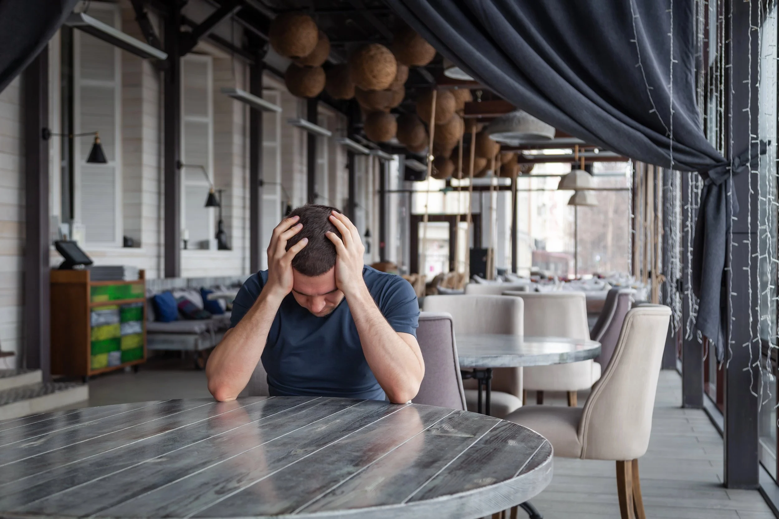 Picture of a man with his head in his hands looking down sitting at a table in an empty roomm