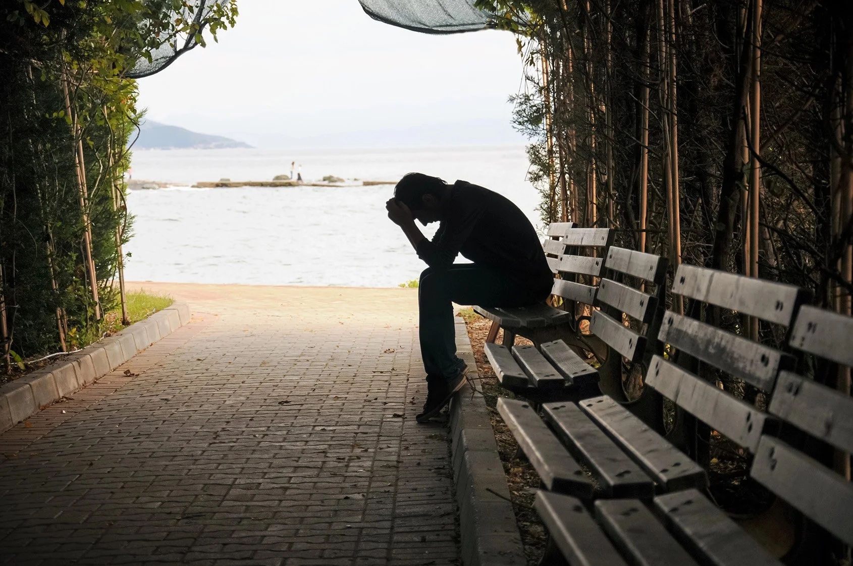 Man sitting on a park bench with his head in his hands in front of a large lake.