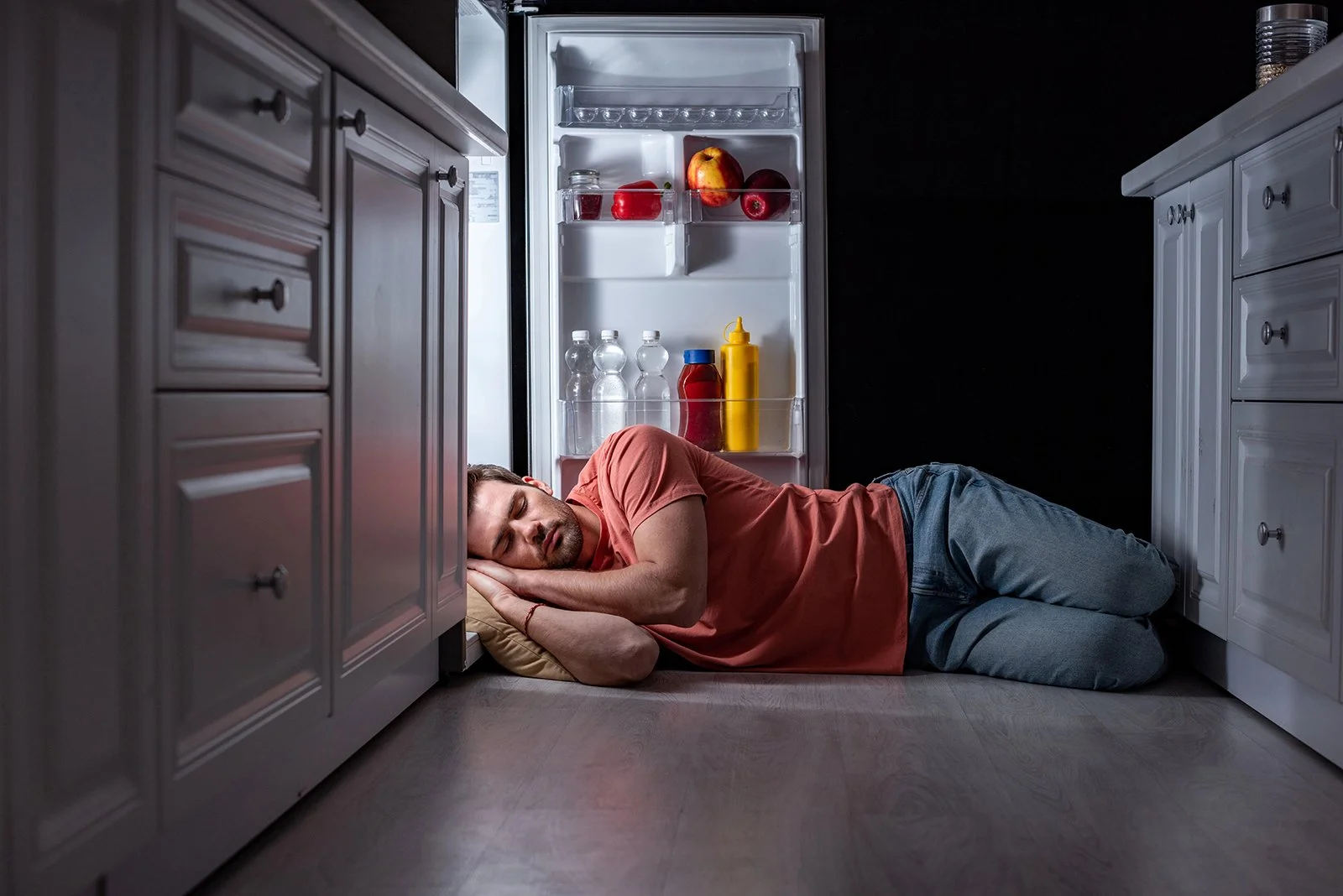 A picture of a man in a a red t-shirt and jeans sleeping on the floor in front of an open refrigerator in a kitchen