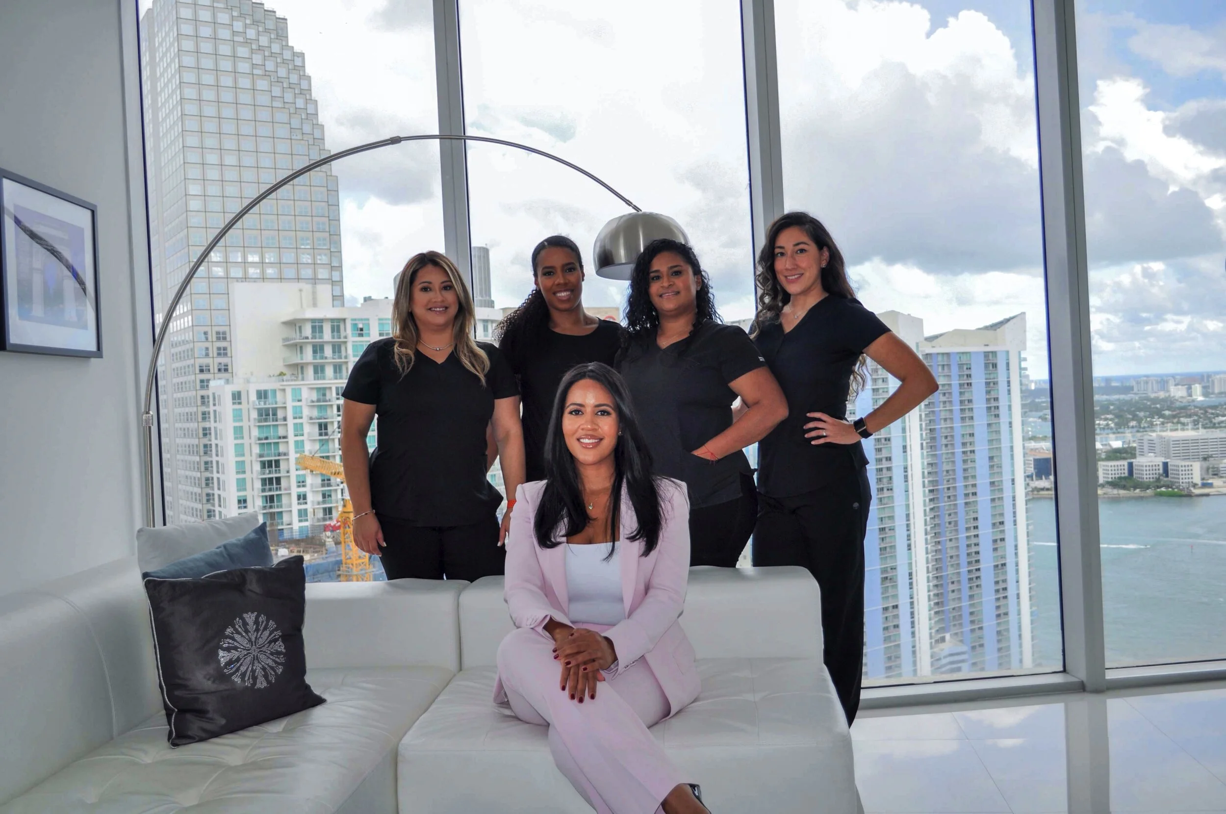 A woman in a pink suit sitting on a white sofa with five women standing behind her in black scrubs, in a high-rise office with city buildings and water view through large windows.