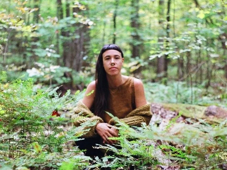 A woman with long dark hair sitting on a mossy log in a dense forest with green foliage.