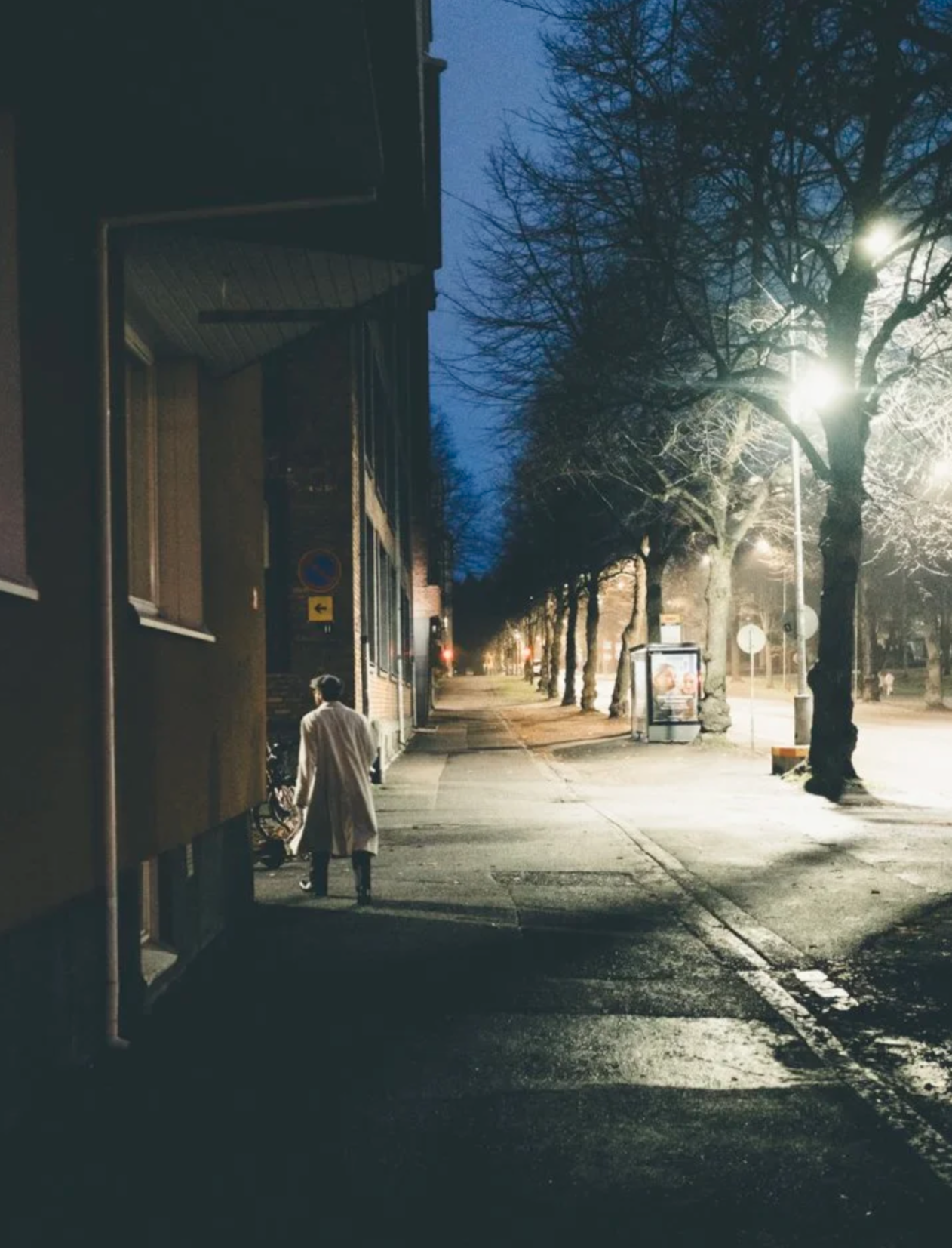 A man walking alone on a sidewalk at night, illuminated by streetlights, with leafless trees lining the street and a building on the corner.