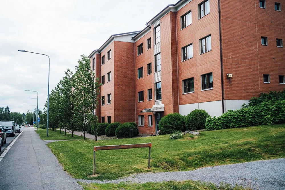 A red brick apartment building with multiple floors, surrounded by green bushes and trees, along a sidewalk with parked cars nearby.