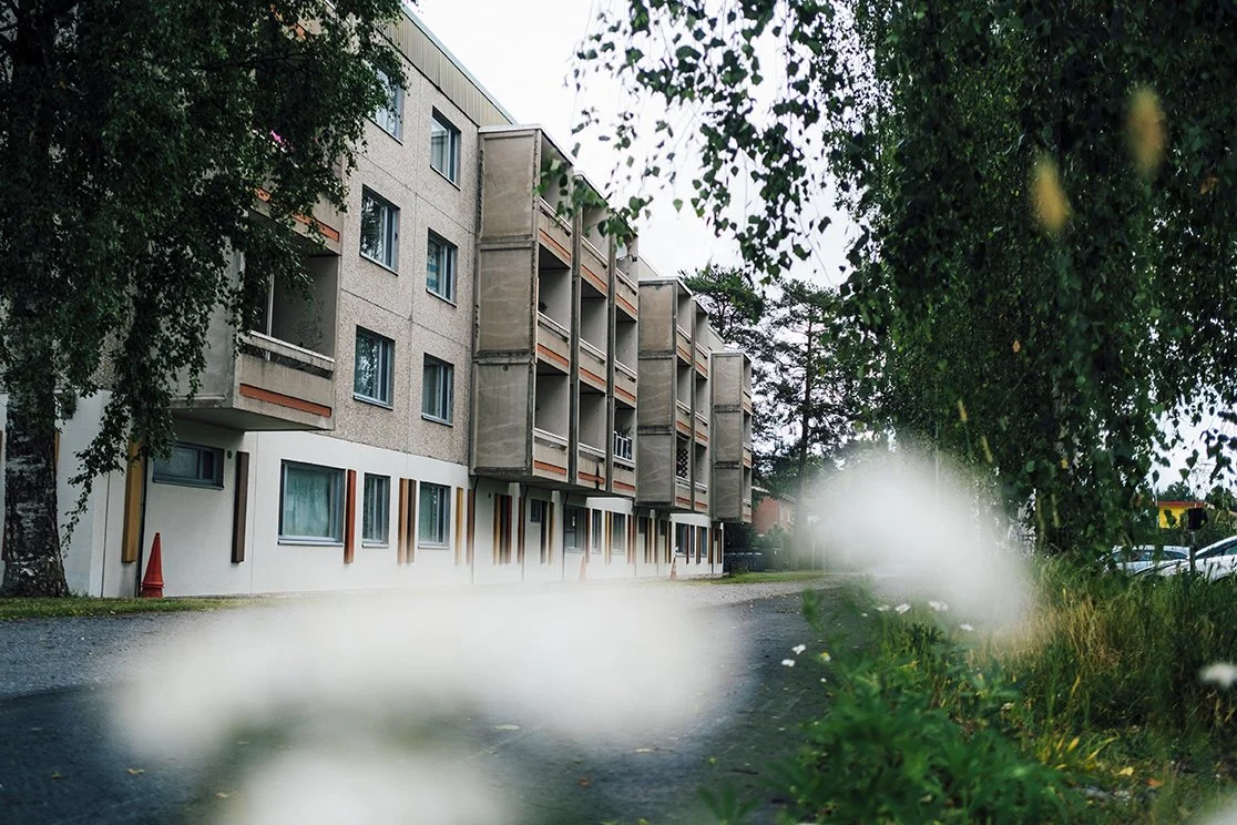 An apartment building with multiple floors, surrounded by green trees and bushes, with an asphalt pathway in front.