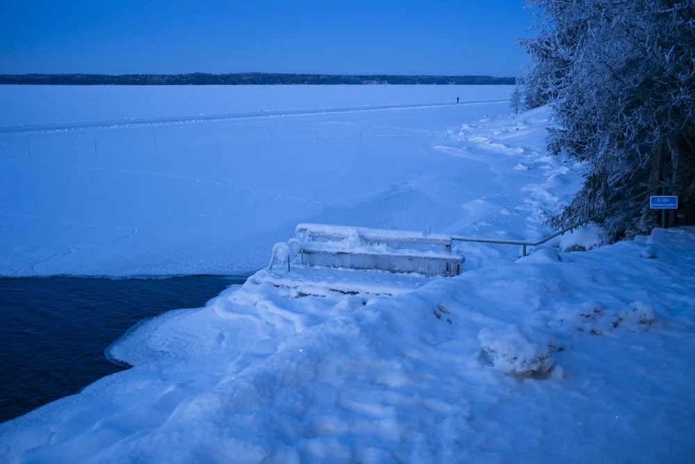 Frozen lake with snow-covered shoreline and stairs leading into the icy water. Snow and ice-covered trees on the right side, with a distant figure in the background.