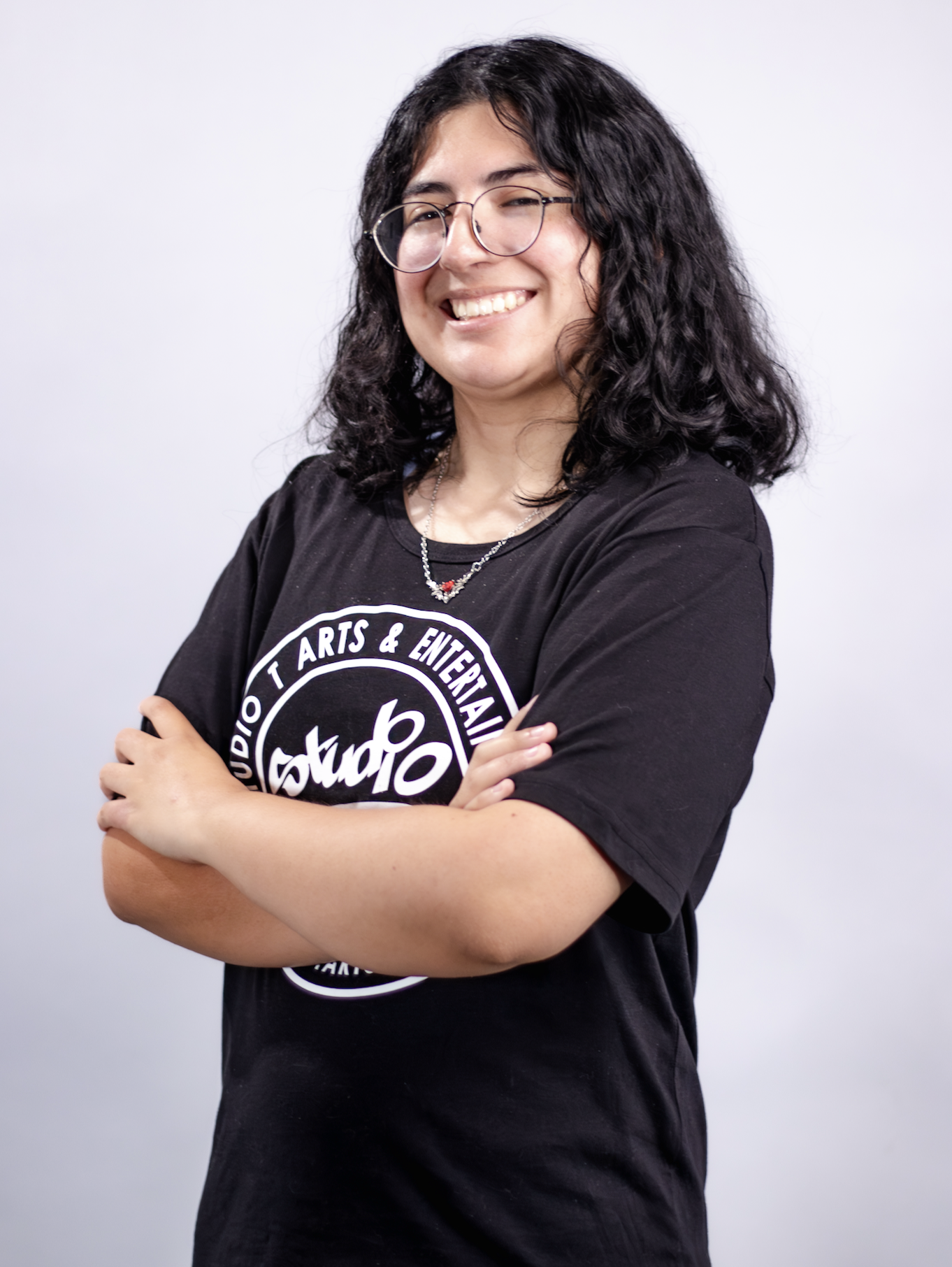 A woman with curly black hair wearing glasses and a black T-shirt with white text, smiling with her arms crossed against a plain white background.