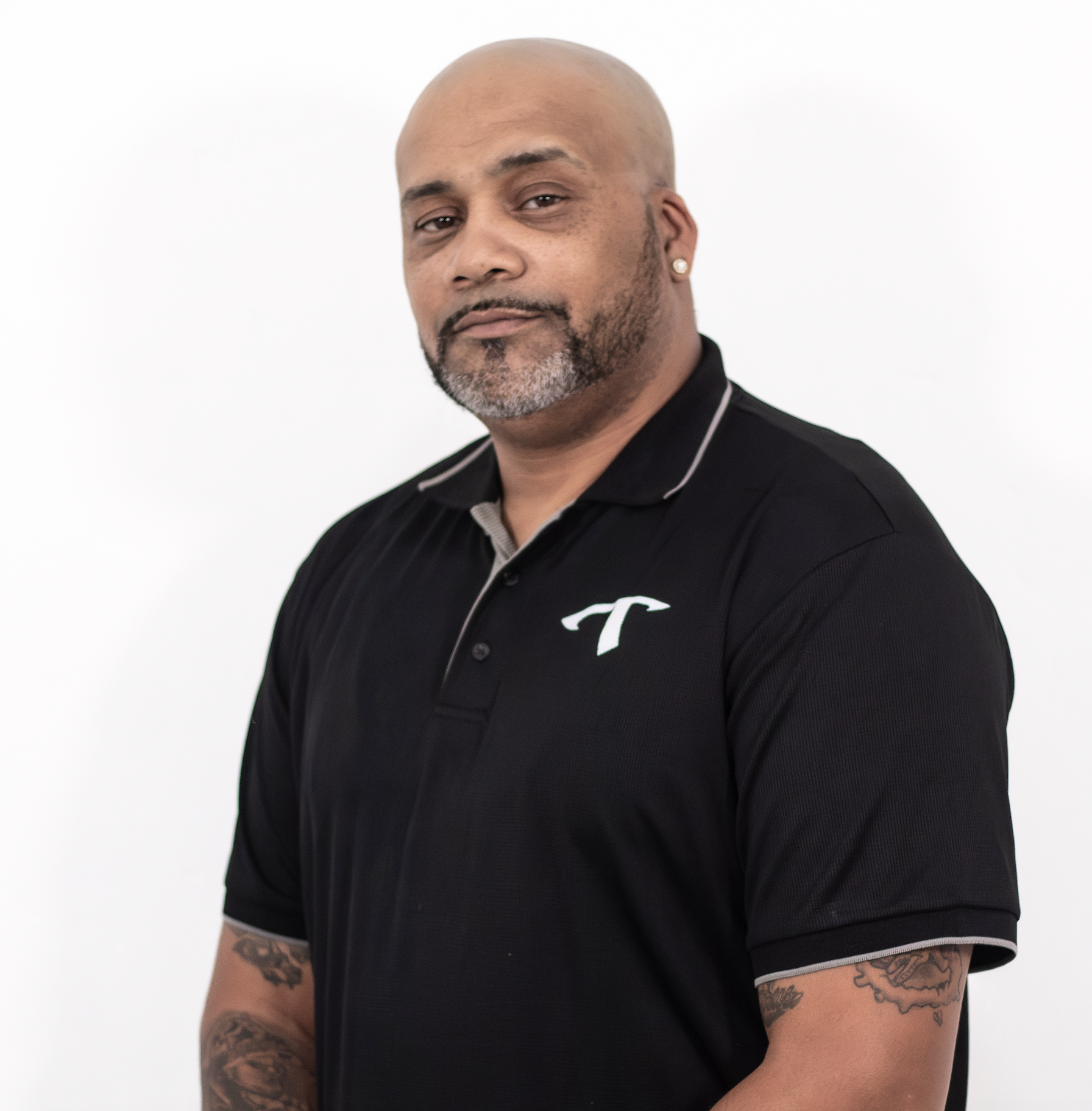Portrait of a bald man with a beard and tattoos on his arms, wearing a black polo shirt with white trim, standing against a plain white background.