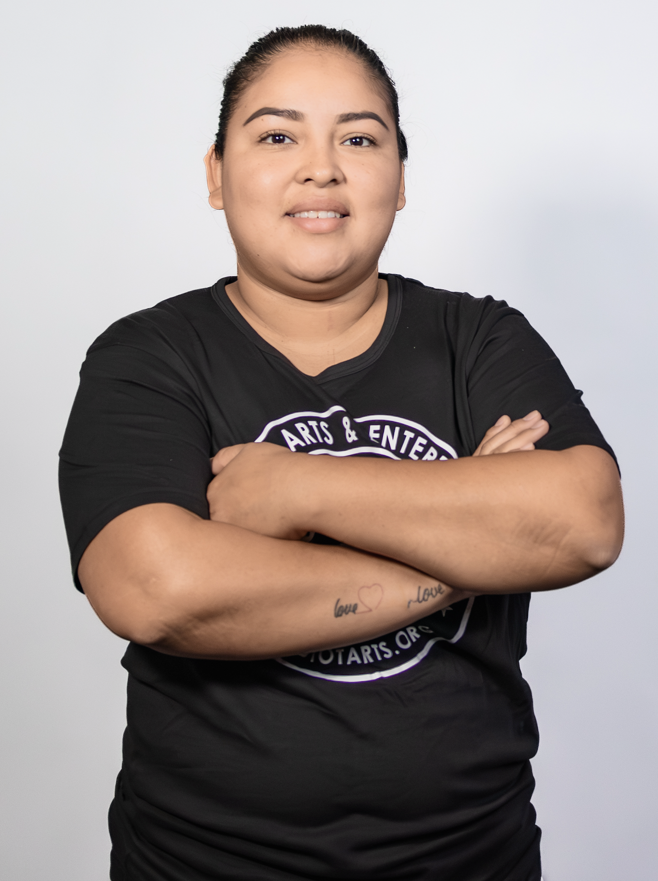 A woman with crossed arms, wearing a black t-shirt that promotes arts and entertainment, standing against a plain white background.