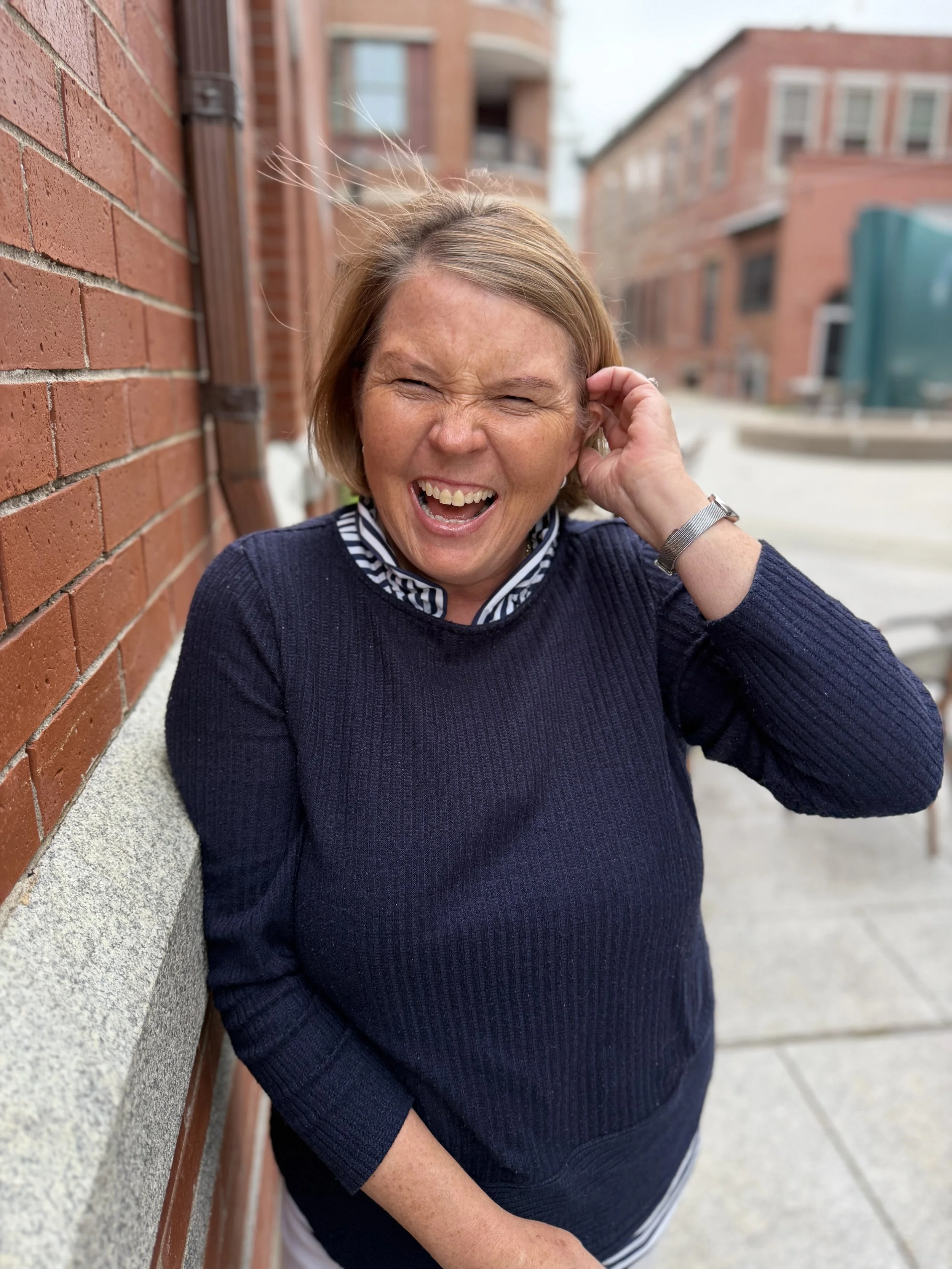 A woman with short brown hair and freckles laughing with her eyes closed, touching her ear, sitting against a brick wall in an urban setting.