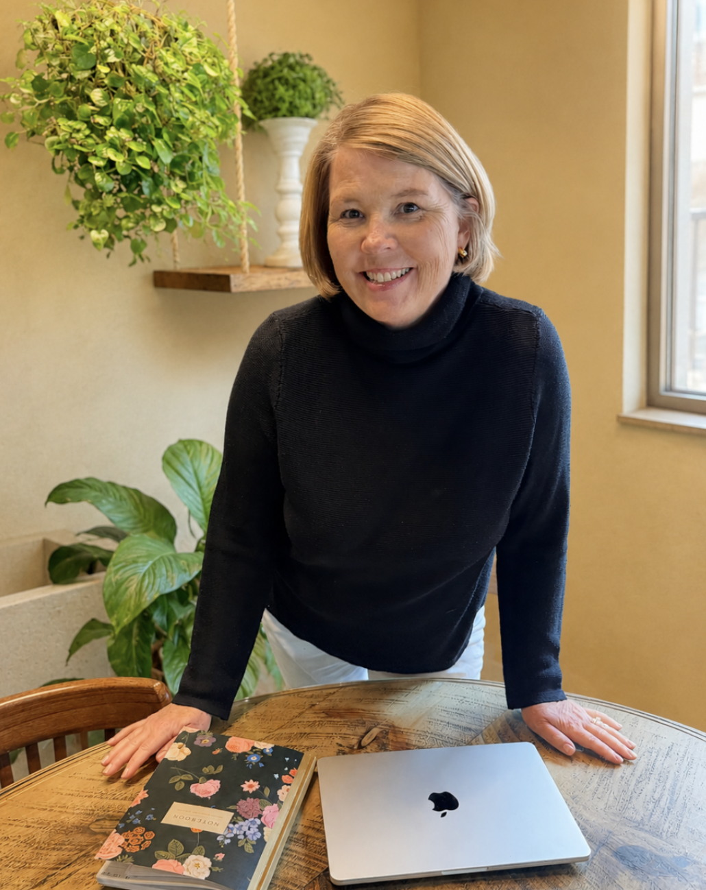 A woman with short blonde hair, wearing a black turtleneck sweater, leaning forward on a wooden table with a closed laptop and a floral notebook. Behind her are green plants and a window letting in natural light.