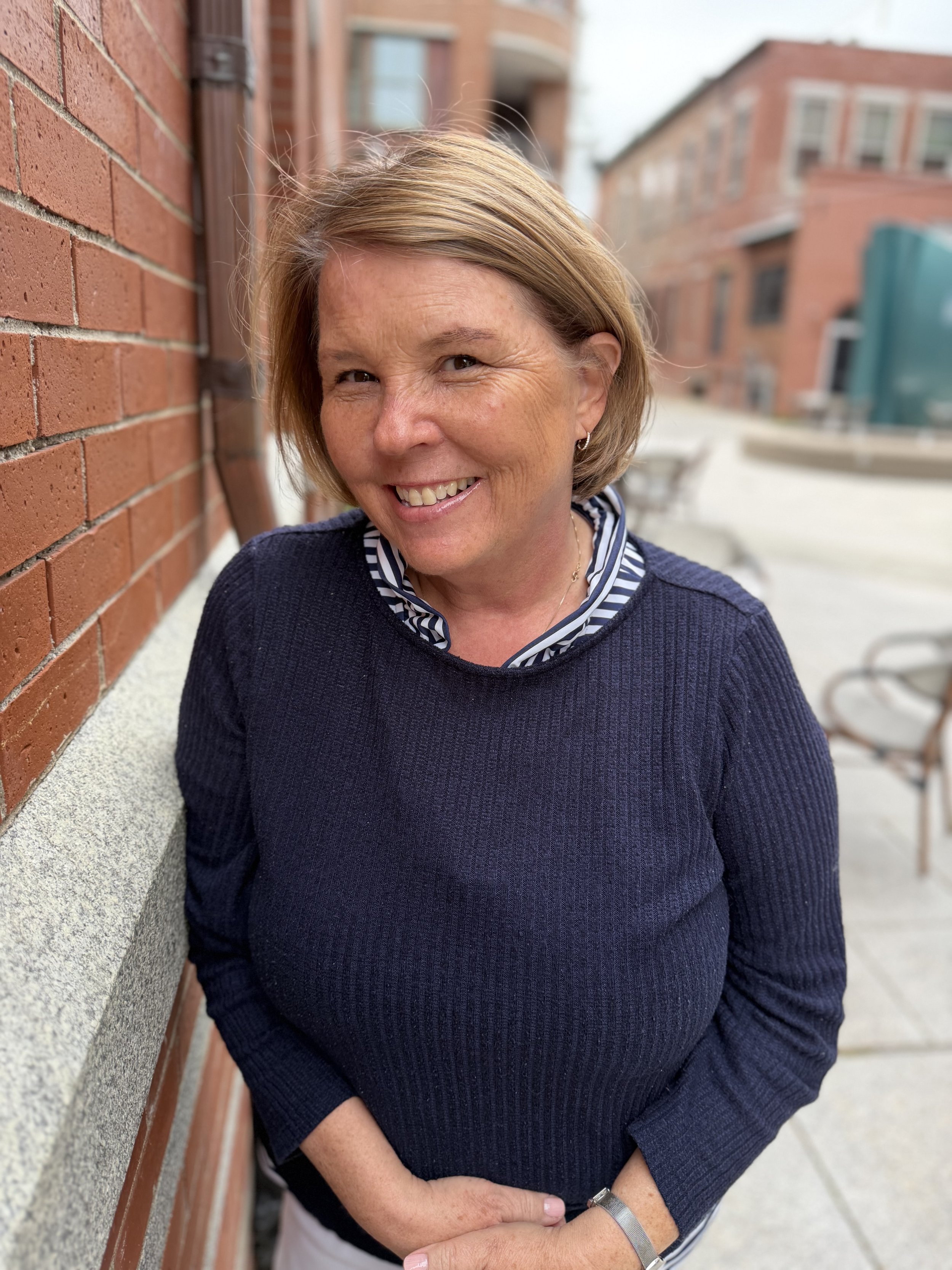 A woman with short blonde hair, smiling, wearing a navy blue sweater, a striped shirt, and a watch, standing outside near a brick wall with a blurred background of buildings and outdoor furniture.