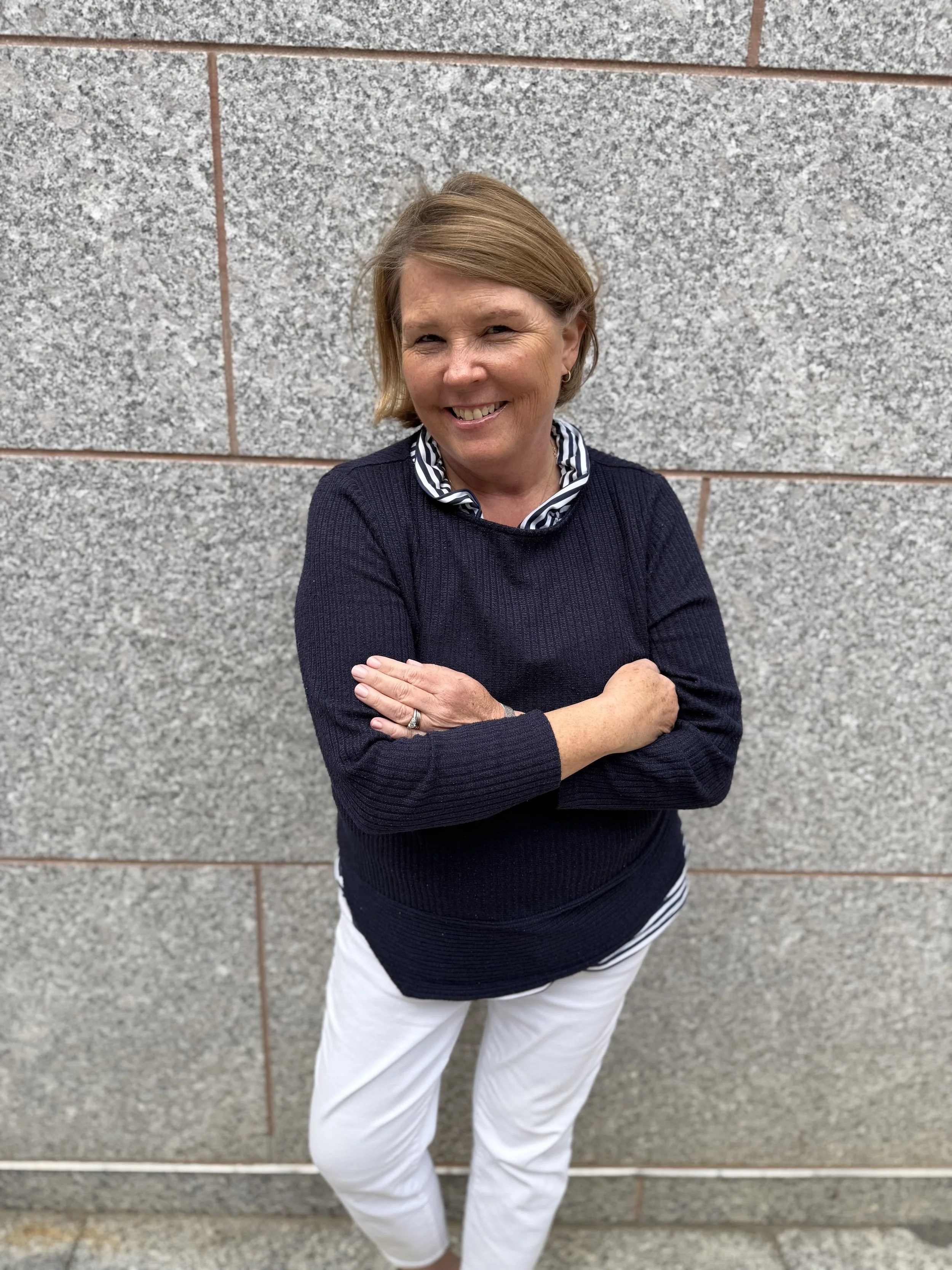 A woman with short blonde hair wearing a navy blue sweater over a striped shirt, standing against a gray stone wall, smiling, with her arms crossed and looking at the camera.