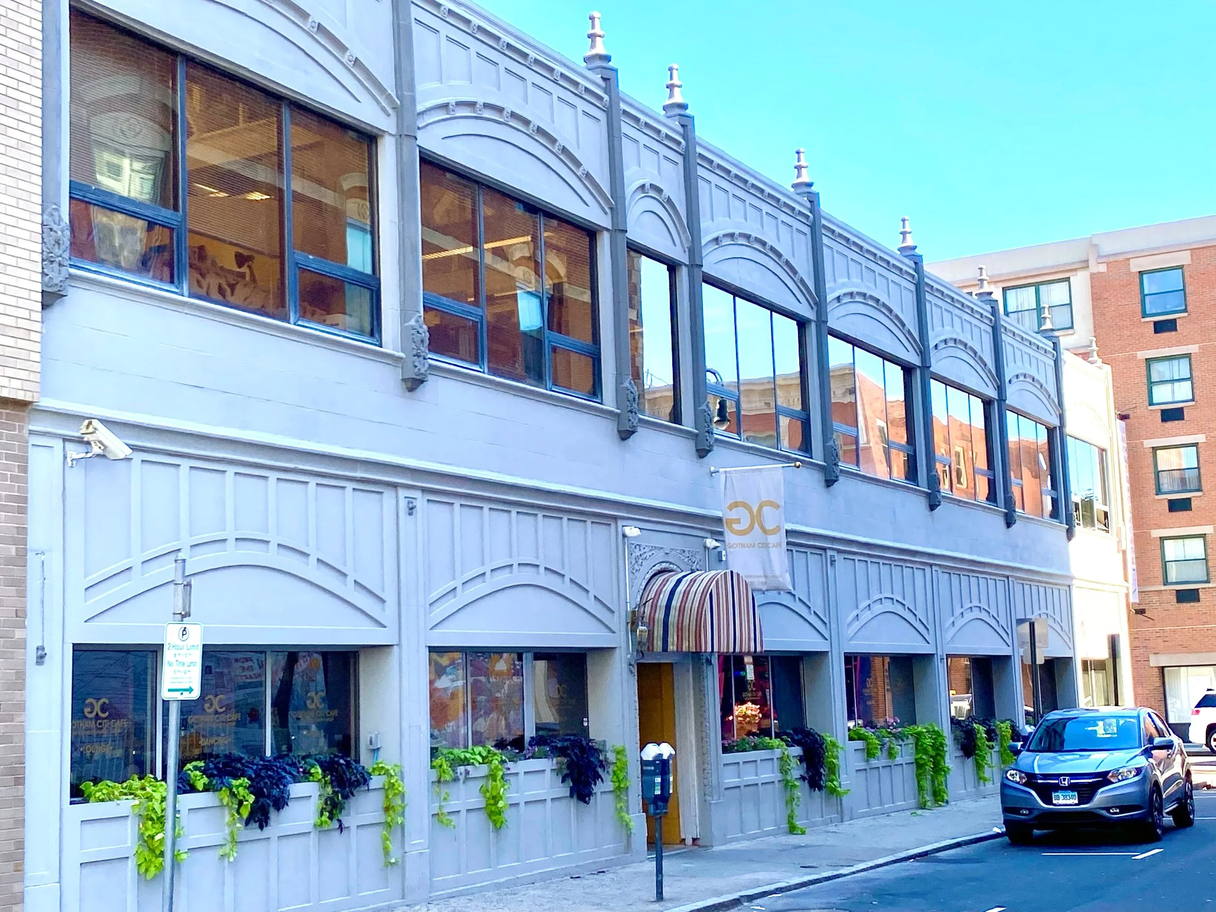 Street view of a two-story building with architectural details, large windows, a striped awning over the entrance, and green plants along the base. A parked car is visible in front.