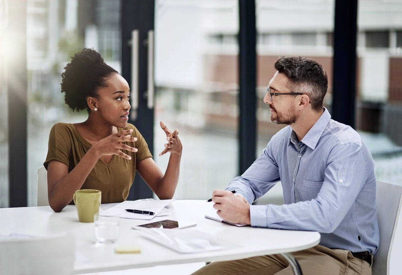 Deux personnes discutent lors d'une réunion à une table, une femme noire gesticulant avec expression, un homme blanc écoutant, dans un cadre professionnel avec grande fenêtre montrant un bâtiment en arrière-plan.