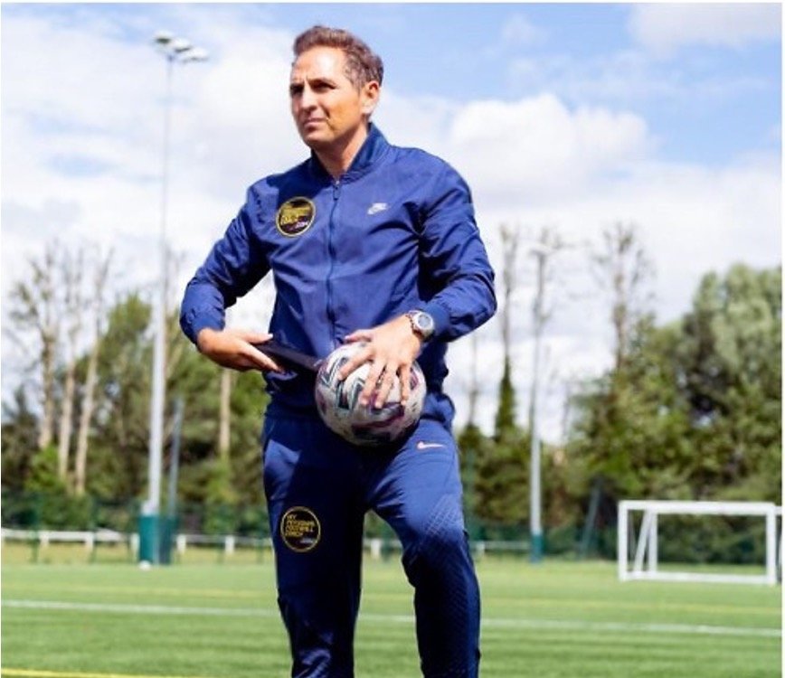 A coach in a blue tracksuit standing on a football field holding a ball and tablet, with trees and a goalpost in the background under a partly cloudy sky.