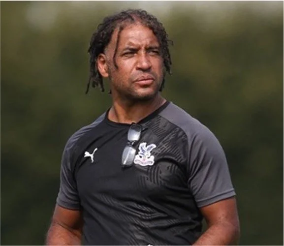 A football coach wearing a black and gray sports shirt with a Puma logo and a crest, standing outdoors with blurred greenery in the background.
