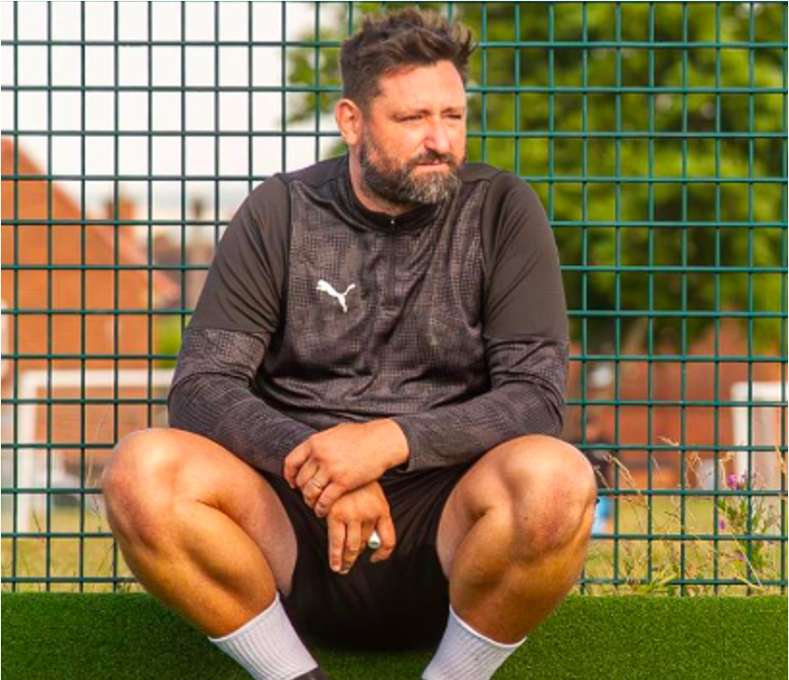 A football coach with beard and dark hair sitting on the grass in front of a green wire fence, wearing black athletic gear and white socks, with residential houses in the background.