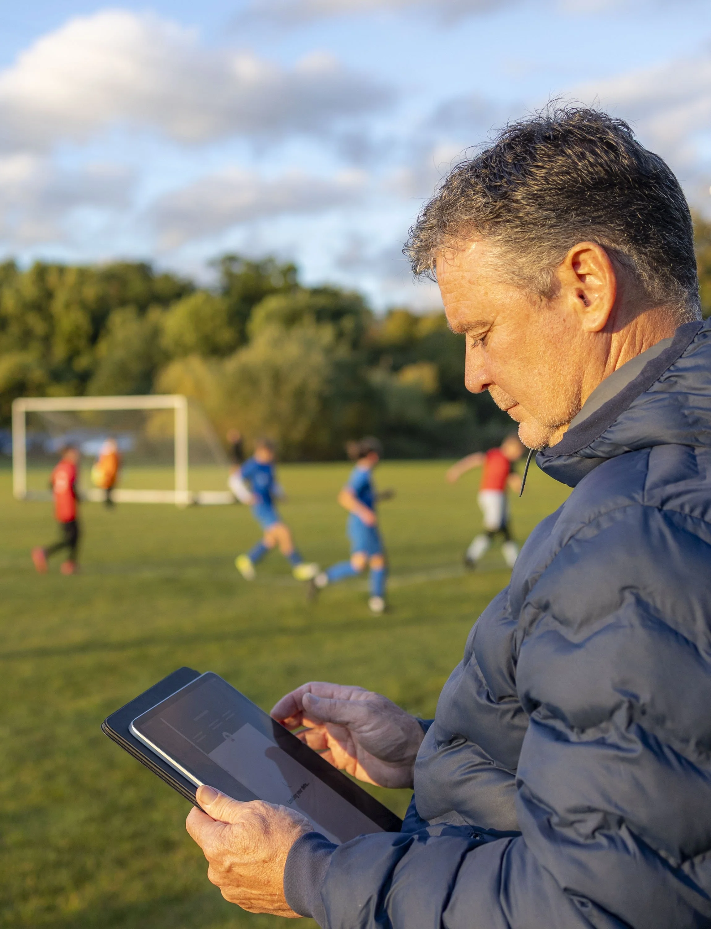 A man in a gray jacket stands on a soccer field holding a tablet, with children playing soccer in the background on a sunny day.