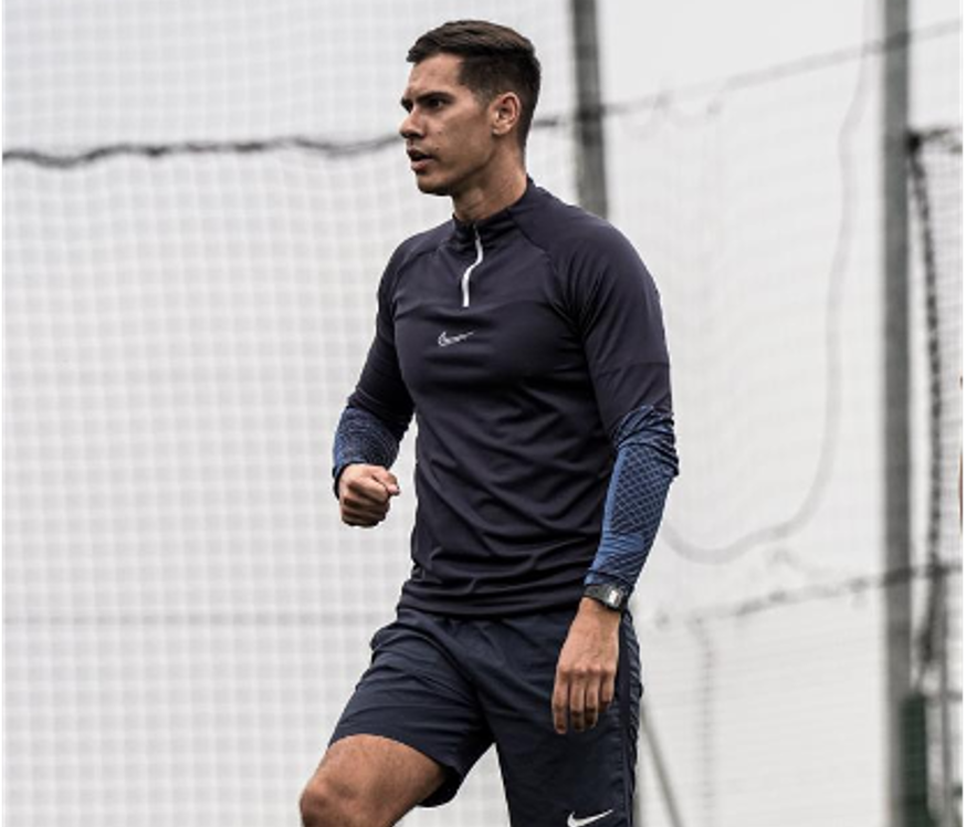 A football coach jogging outdoors near a mesh fence, wearing a black Nike athletic jacket and shorts, with a watch on his left wrist.