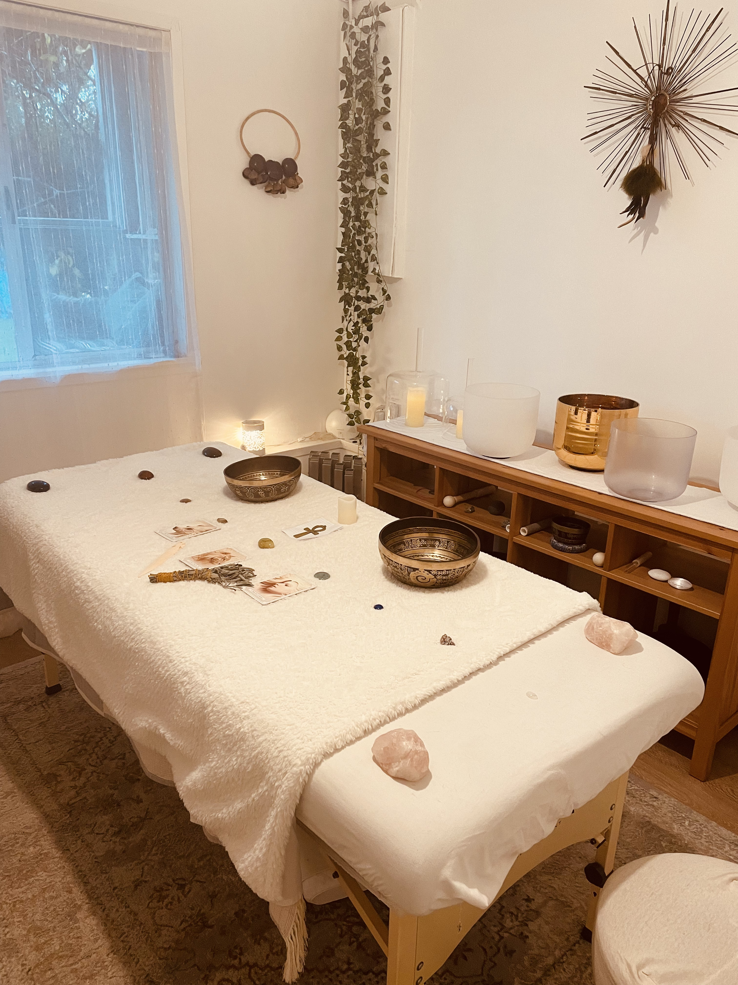 A massage or wellness room with a massage table covered in a white blanket, decorated with crystals, stones, candles, and tarot cards. The room has a window with curtains, a wall decorated with hanging plants and a starburst wall art, and contains cr