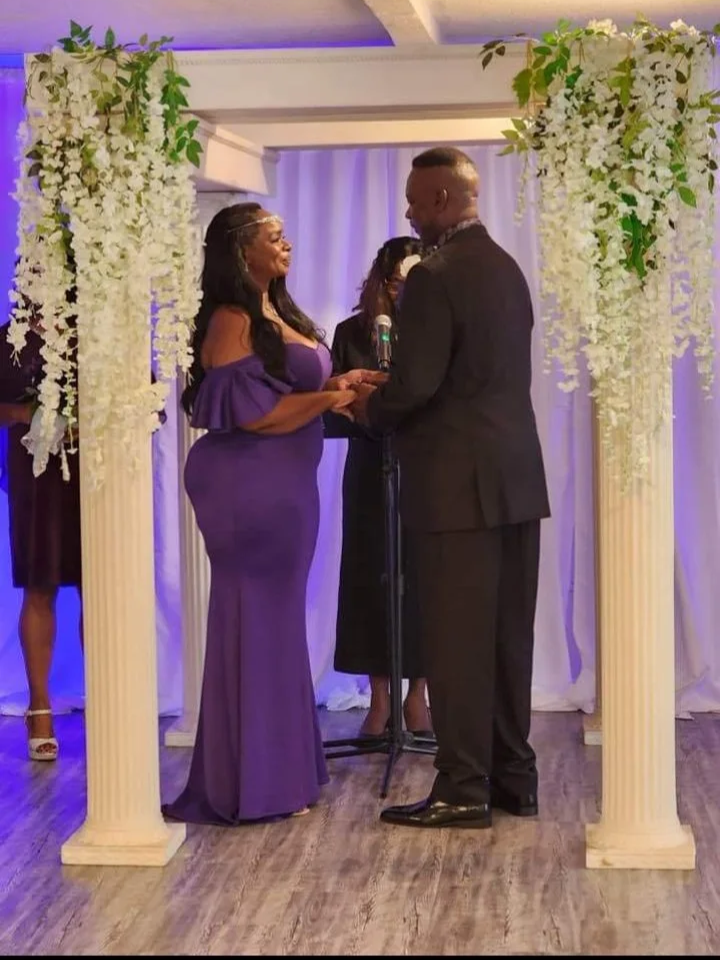 A same-sex couple exchanges vows during a wedding ceremony under a decorated arch with white flowers and greenery, with a officiant present.